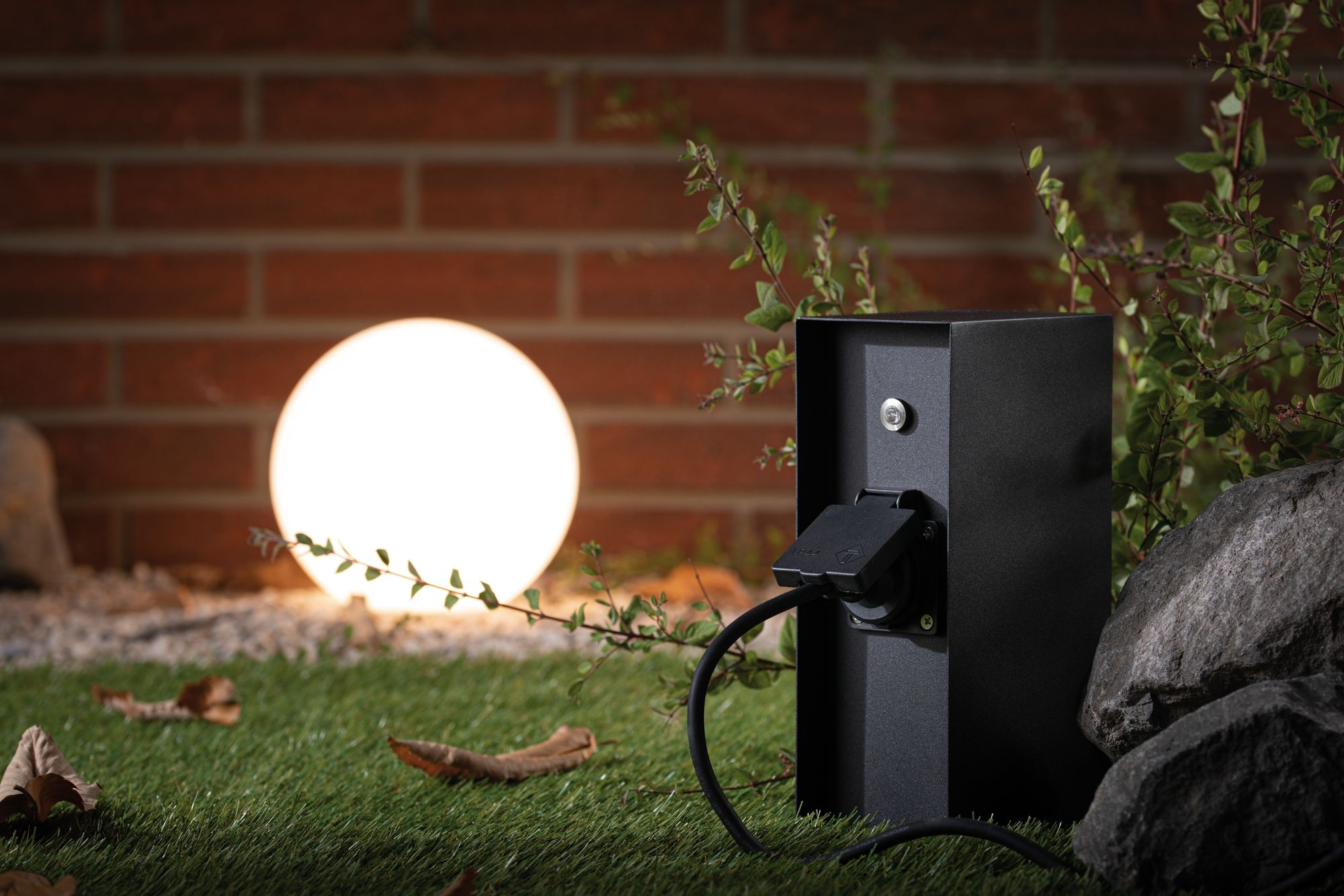 Czarna zewnętrzna skrzynka gniazdka elektrycznego z podłączonym kablem, świecącą kulą i ceglanym murem w tle.