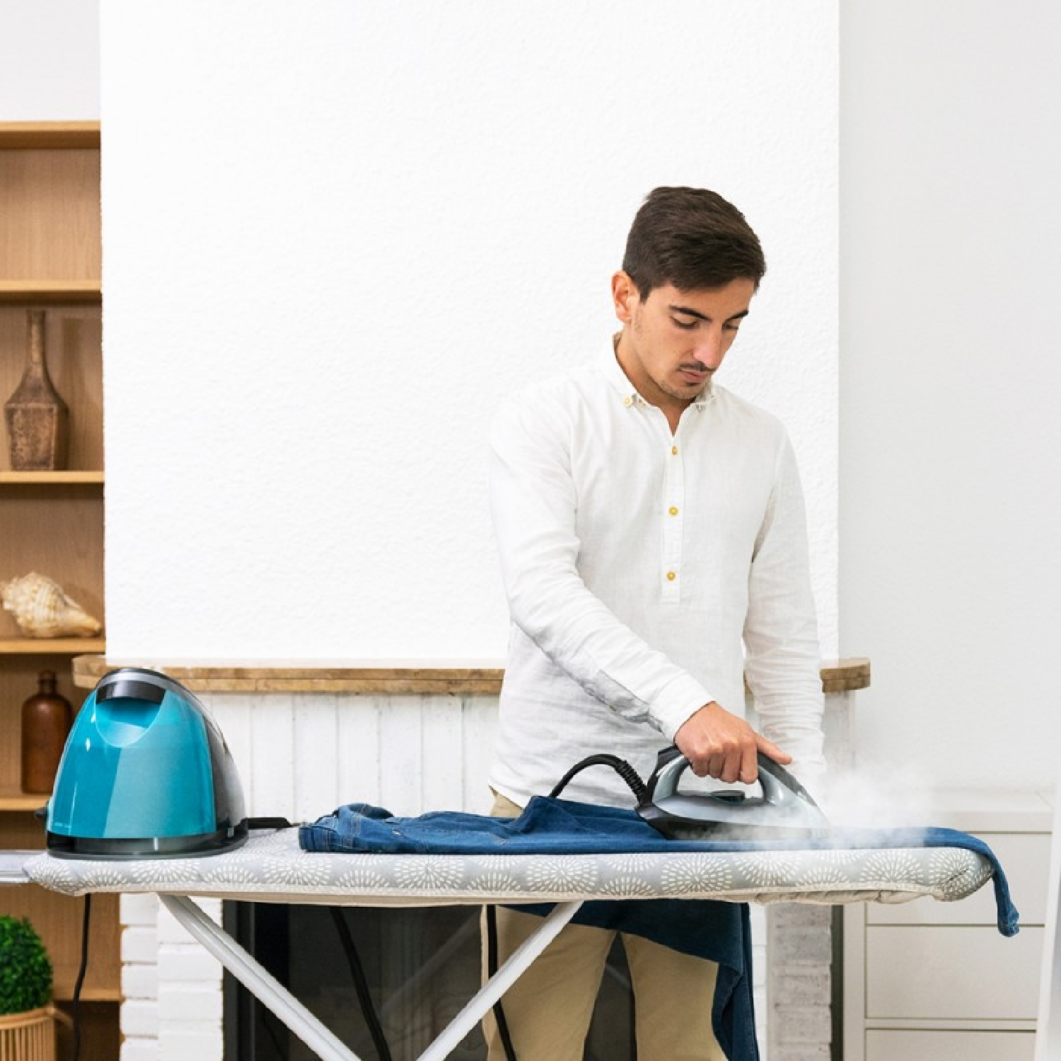 Mann bügelt ein blaues Jeanshemd auf einem Bügelbrett. Dampf ist sichtbar. Weißer Wandhintergrund.