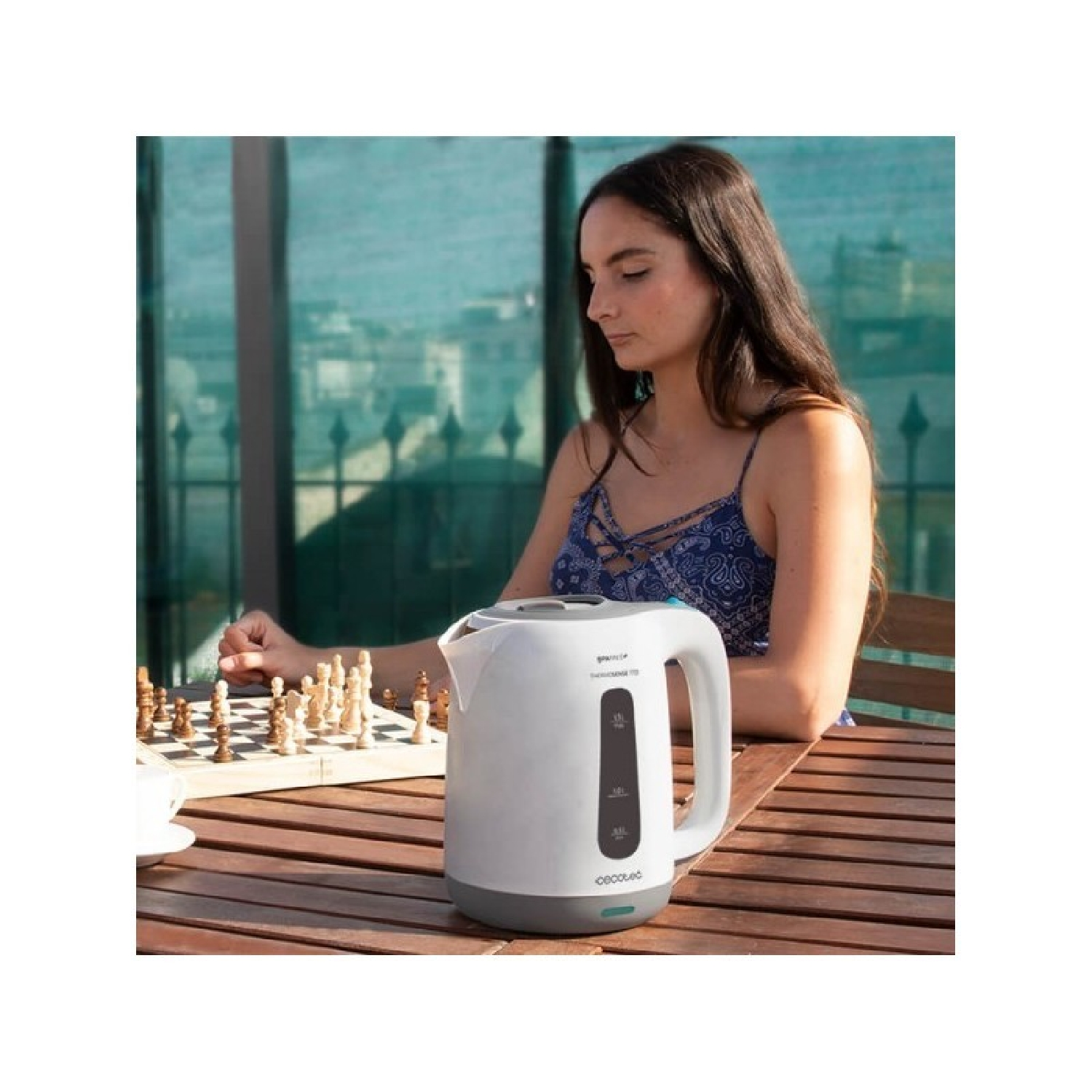 Una donna che gioca a scacchi con un bollitore elettrico bianco sul tavolo. La scena è all'aperto.