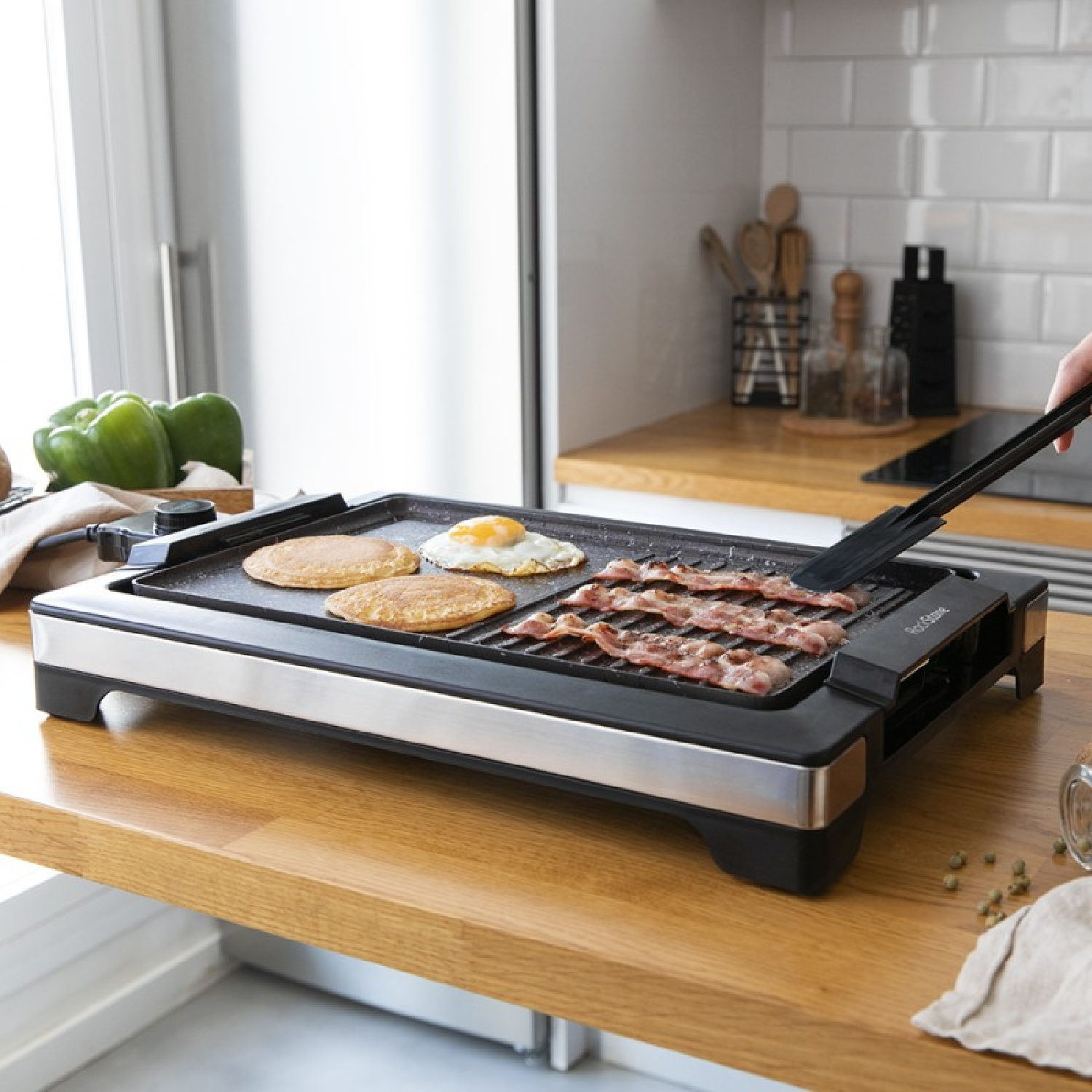 Una piastra elettrica con pancake, uova e pancetta. Una persona usa le pinze per spostare la pancetta.