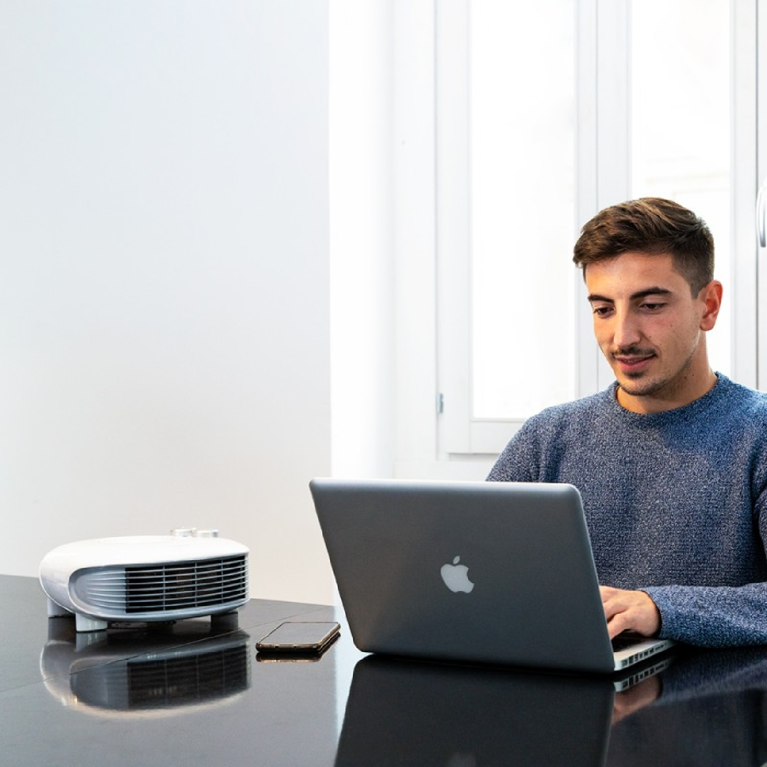 Mężczyzna pracuje na laptopie. Na biurku stoi biały wentylator i telefon. Tło jest białe.
