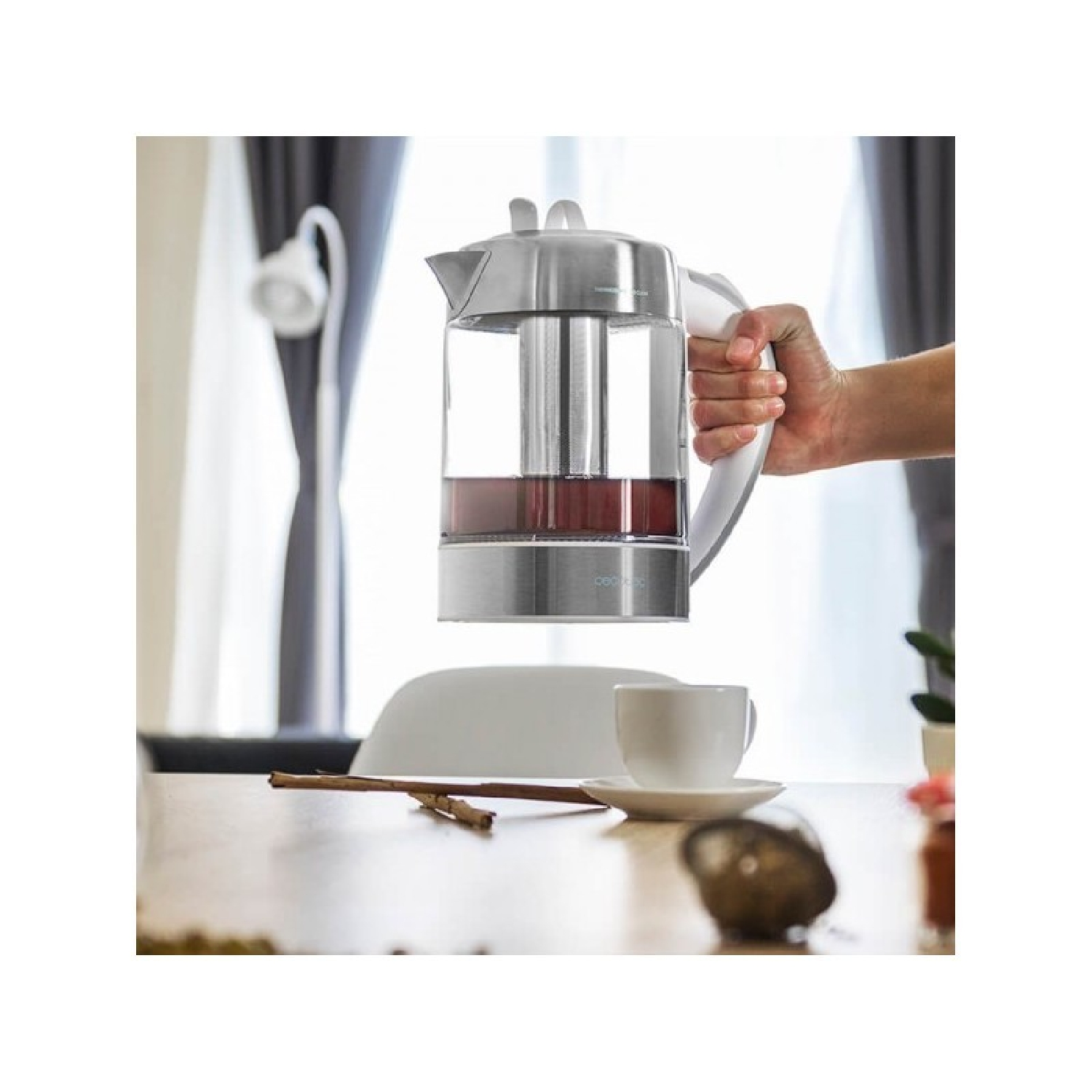Die Hand einer Person, die einen Glasteekessel hält. Tasse und Untertasse auf einem Tisch im Hintergrund.
