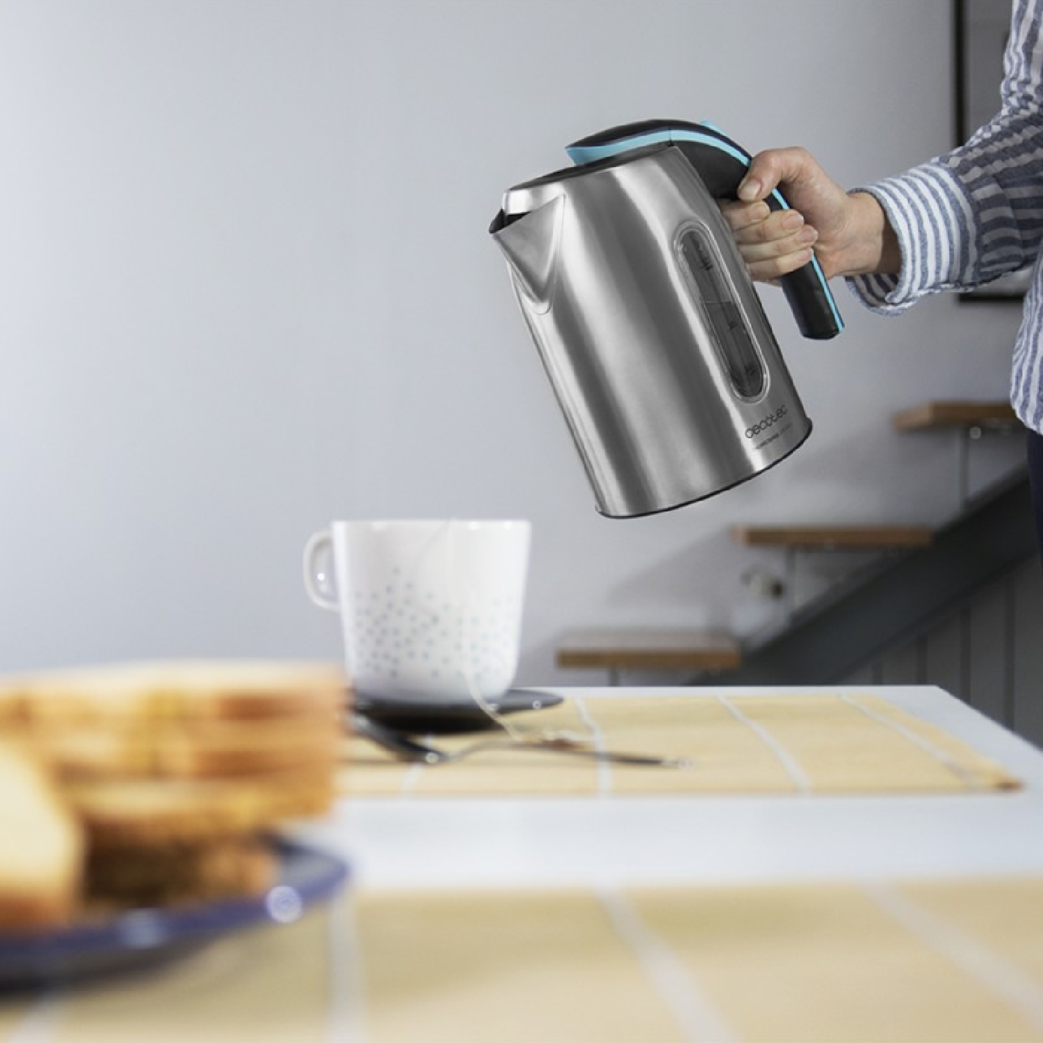 Hervidor de agua plateado que se vierte en una taza blanca sobre una mesa. Pan rebanado en el fondo.