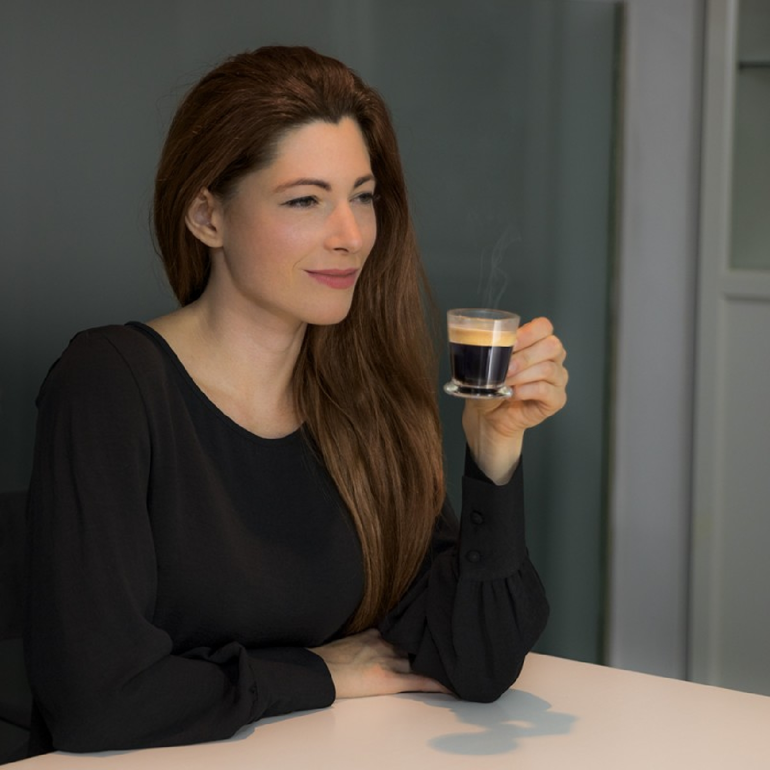Kobieta z długimi brązowymi włosami trzyma małą szklankę kawy, patrząc w bok, w pokoju z białym stołem.