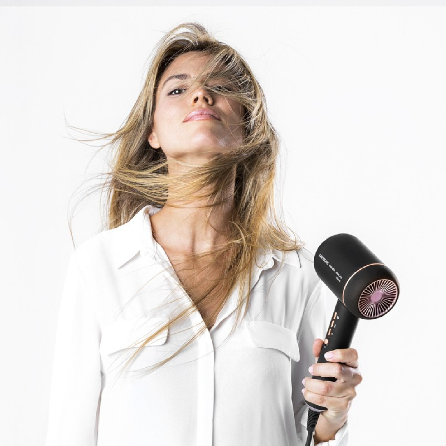 Una donna con i capelli biondi, che tiene un asciugacapelli nero e oro rosa, guarda verso la fotocamera.