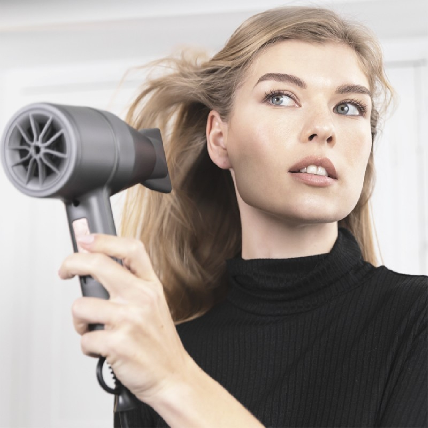 Una donna si asciuga i capelli con un asciugacapelli grigio. Sta guardando in alto. Capelli biondi.