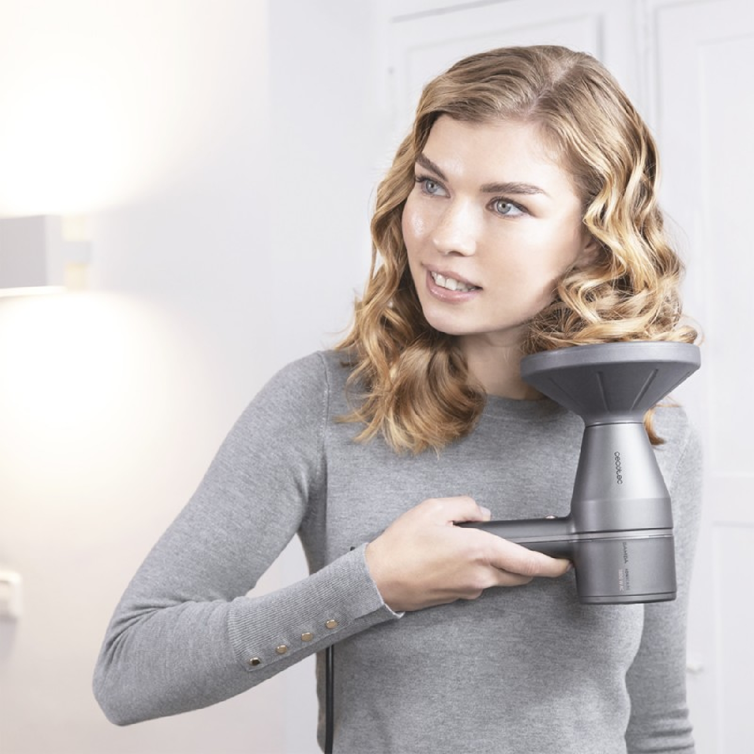 Donna con un asciugacapelli, sorride leggermente. Ha i capelli biondi e si trova in una stanza bianca.