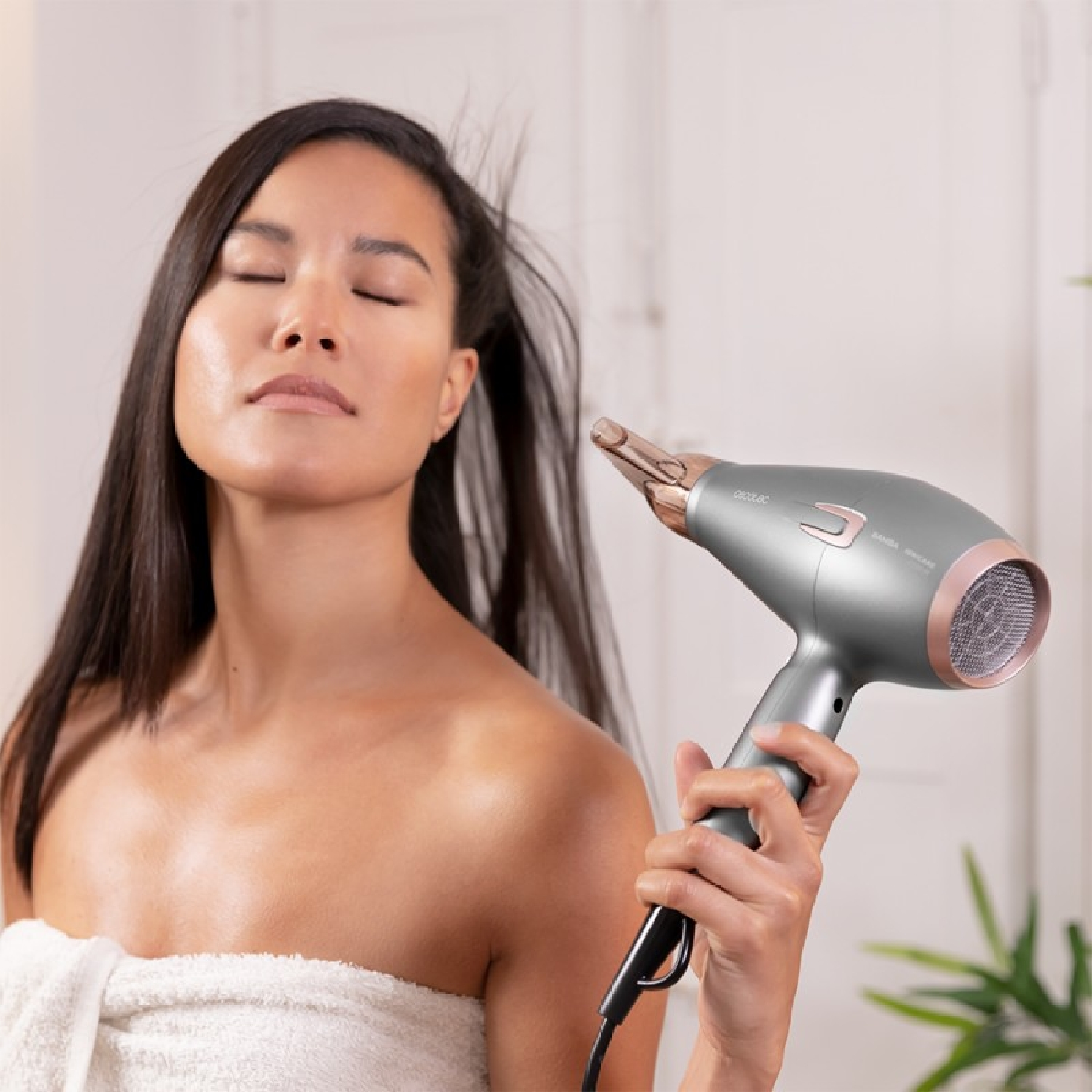 Una donna con lunghi capelli scuri usa un asciugacapelli grigio e oro rosa. Ha un asciugamano e gli occhi chiusi.