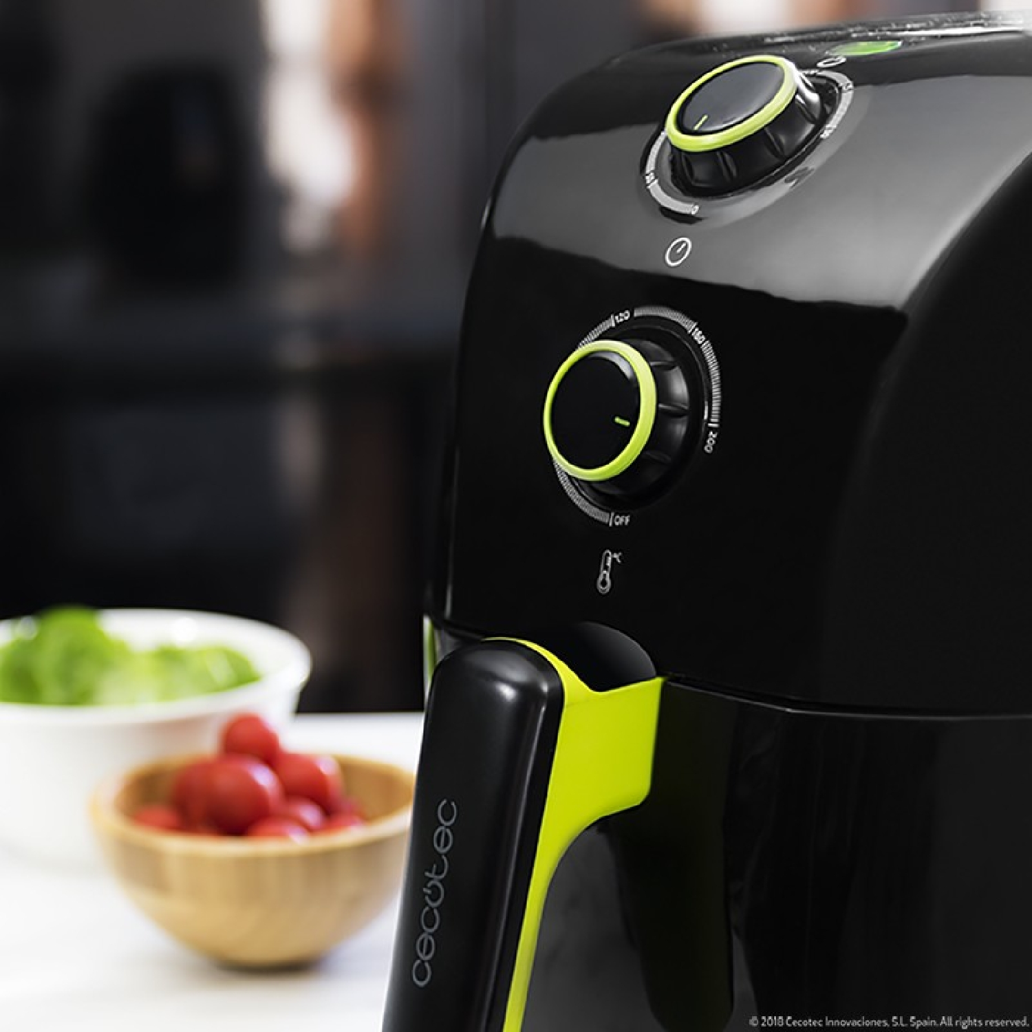 Freidora de aire negra con acentos verdes. Perillas visibles, comida al fondo.