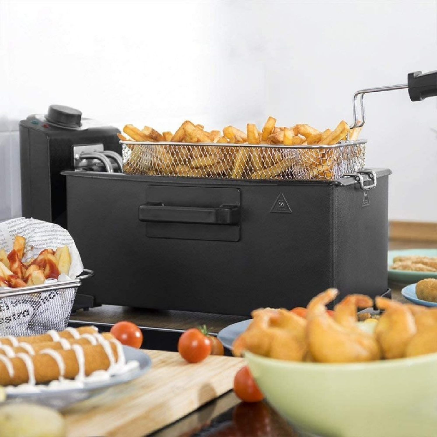 Freidora negra con cesta de patatas fritas. Se ven otros alimentos fritos, tomates y salsa para mojar.