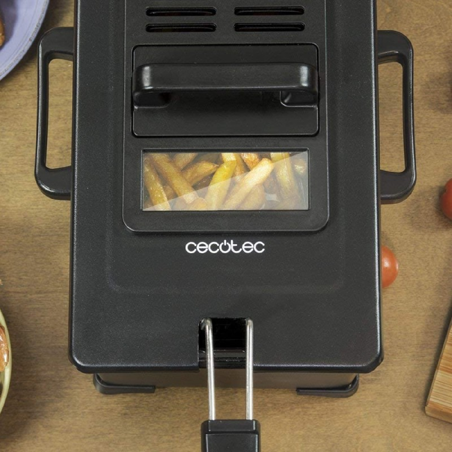 Freidora de aire negra con patatas fritas dentro, colocada sobre una superficie de madera con tomates y tabla de cortar.