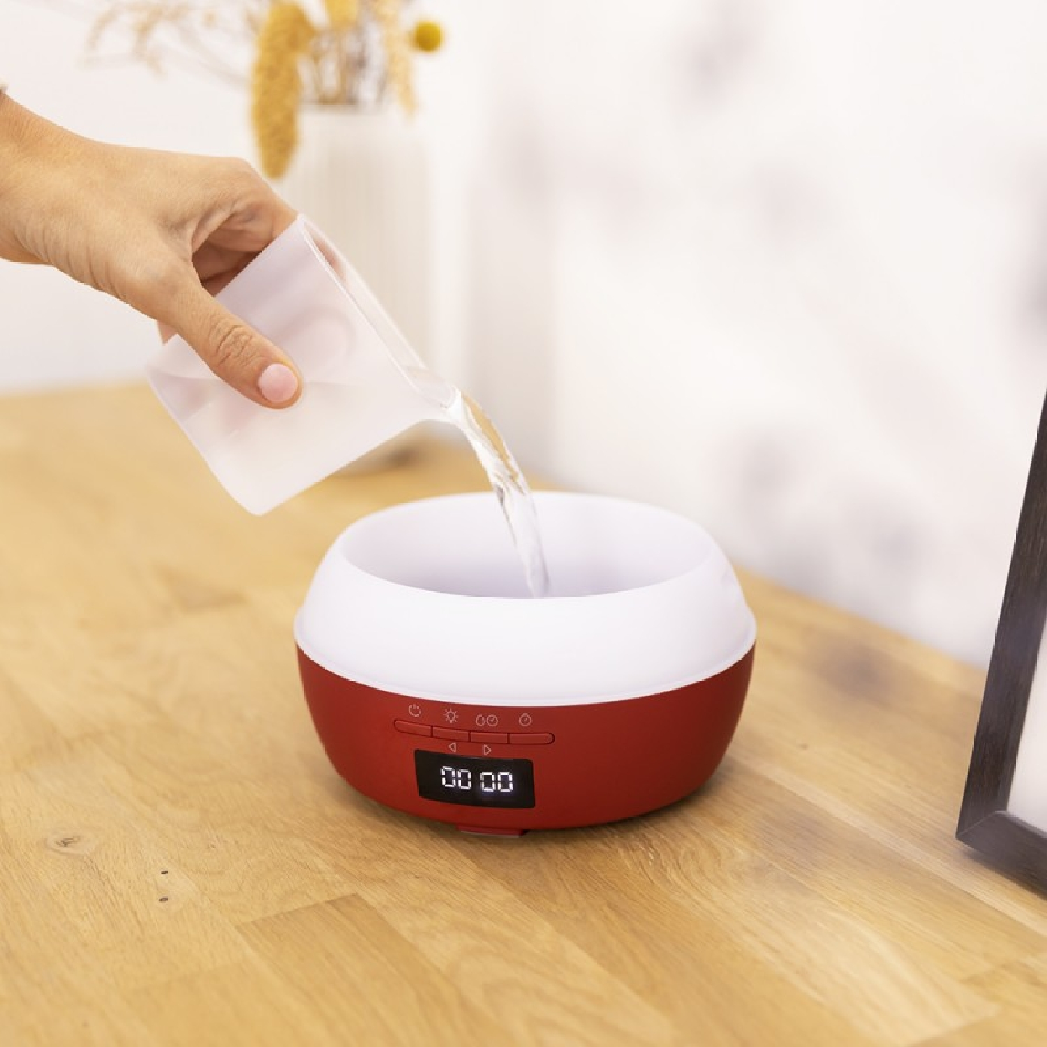 Ręka wlewa wodę do czerwono-białego dyfuzora. Drewniany stół, biały wazon w tle.