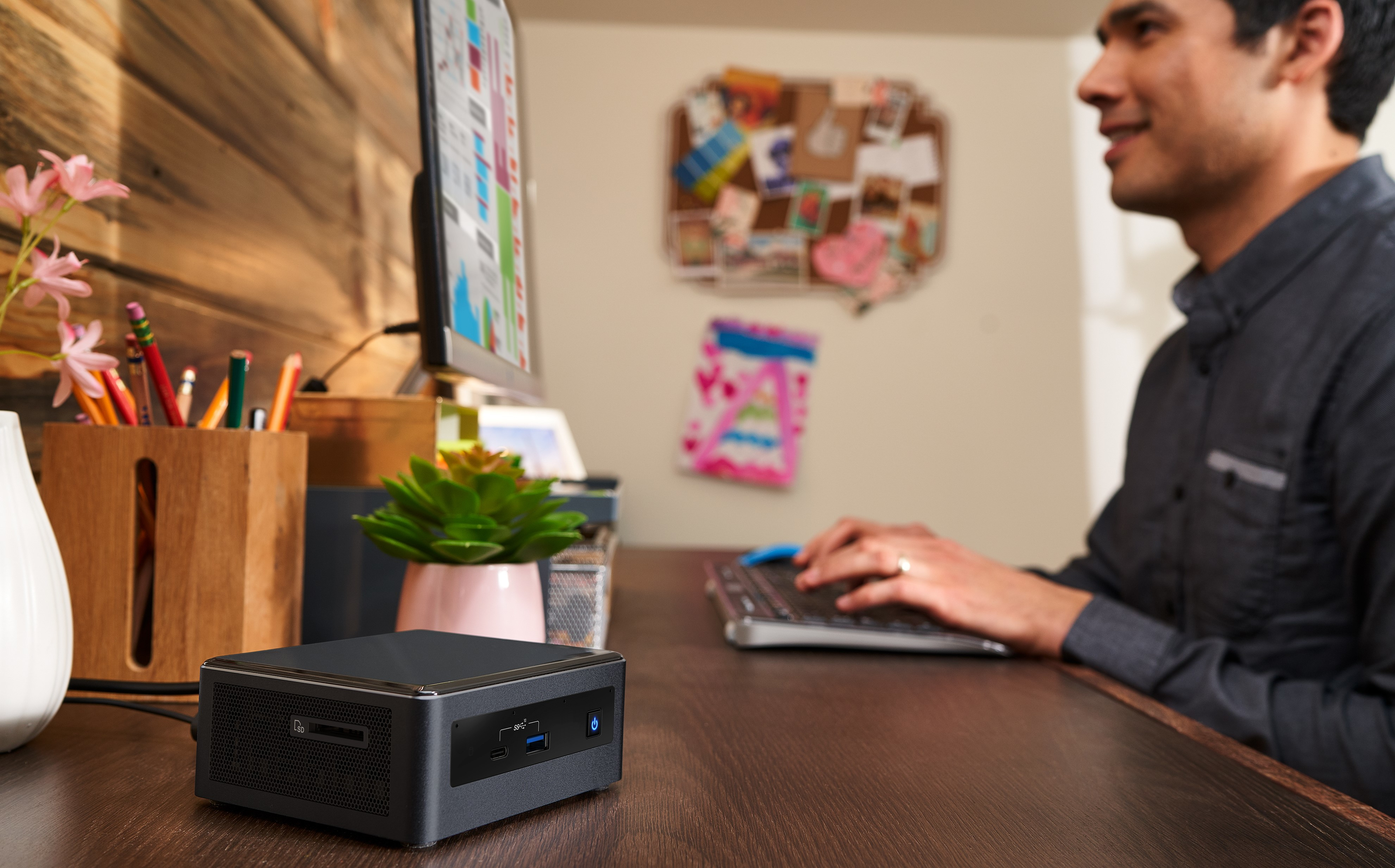 Mann arbeitet an einem Schreibtisch mit Computer. Mini-PC und Pflanzen im Vordergrund.