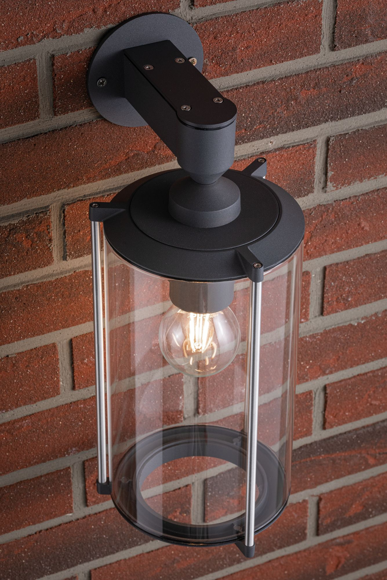 Schwarze Lampe an Backsteinmauer, mit klarem Glasschirm und sichtbarer Glühbirne.