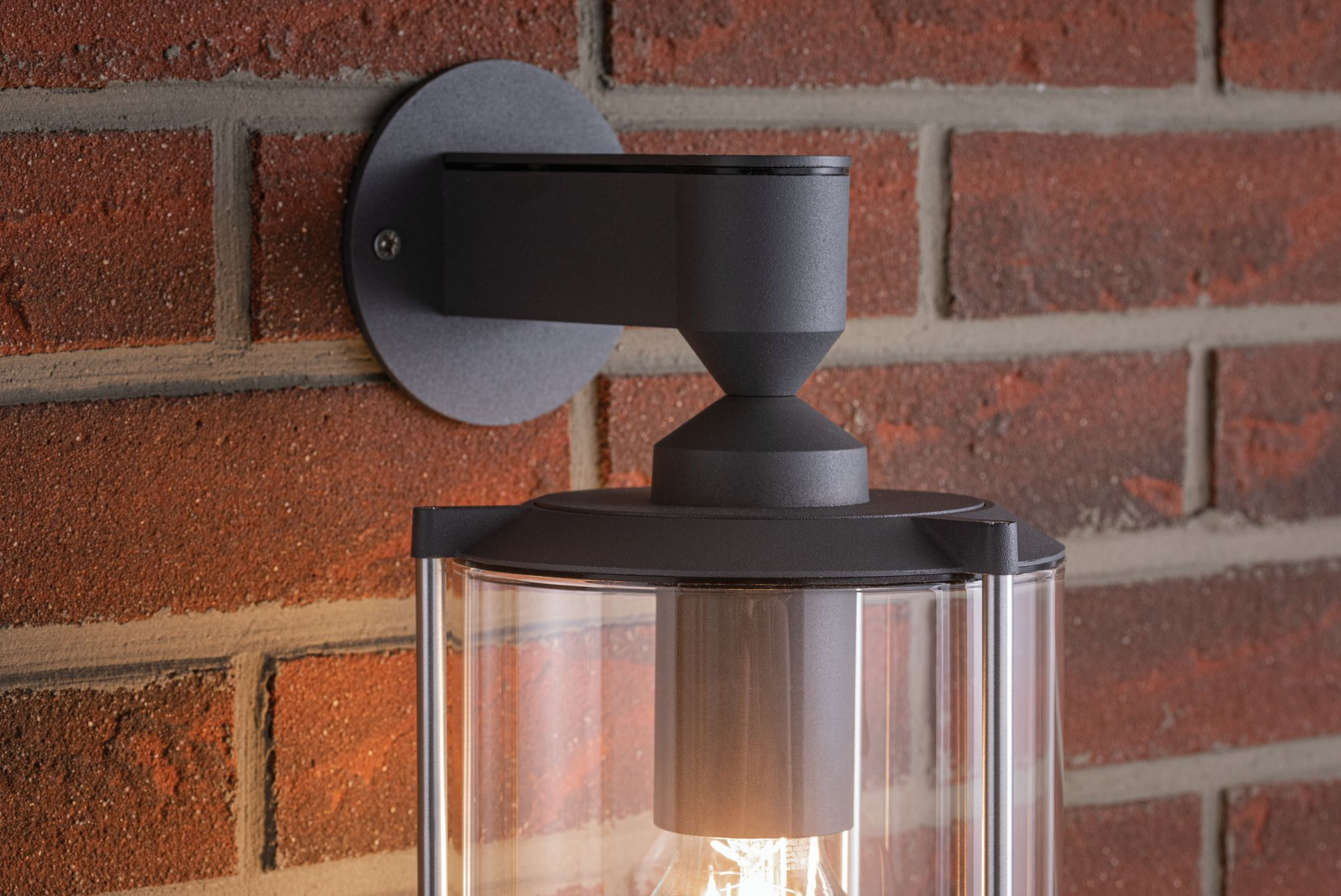 Außenwandleuchte an einer roten Backsteinmauer. Die Lampe hat ein schwarzes Gehäuse und ein Glasgehäuse.