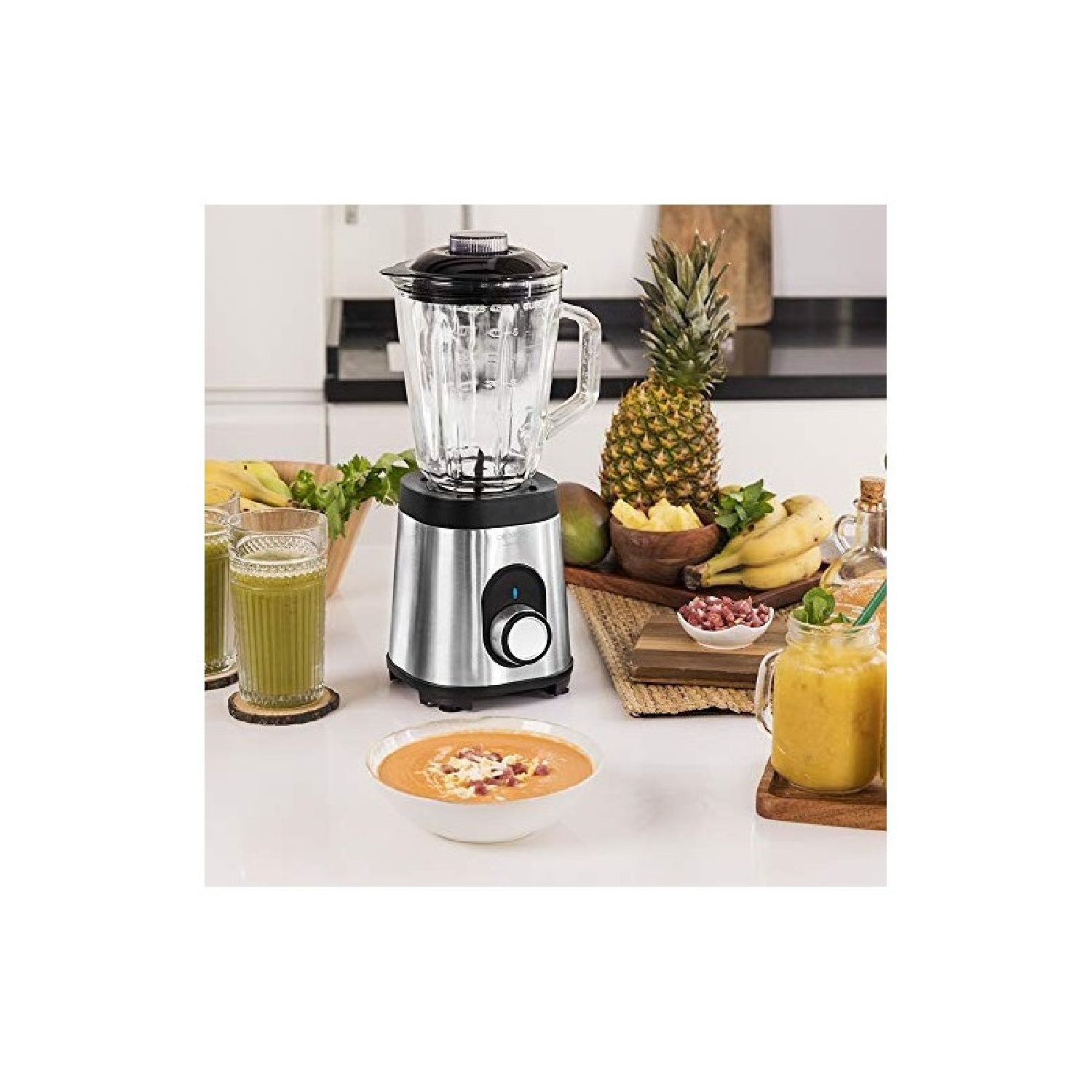 Ein silberner Mixer mit einem Glasbehälter, Früchten und Smoothie-Gläsern auf weißer Oberfläche.