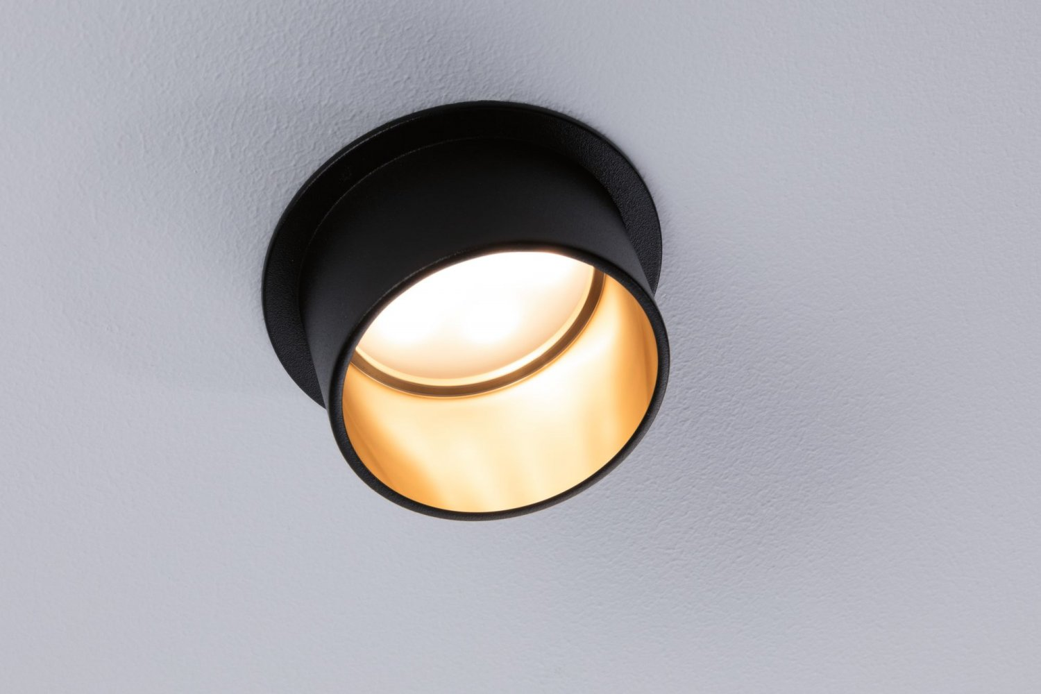 Un encastré noir. La lumière est allumée et brillante à l'intérieur du luminaire. Monté sur un plafond blanc.