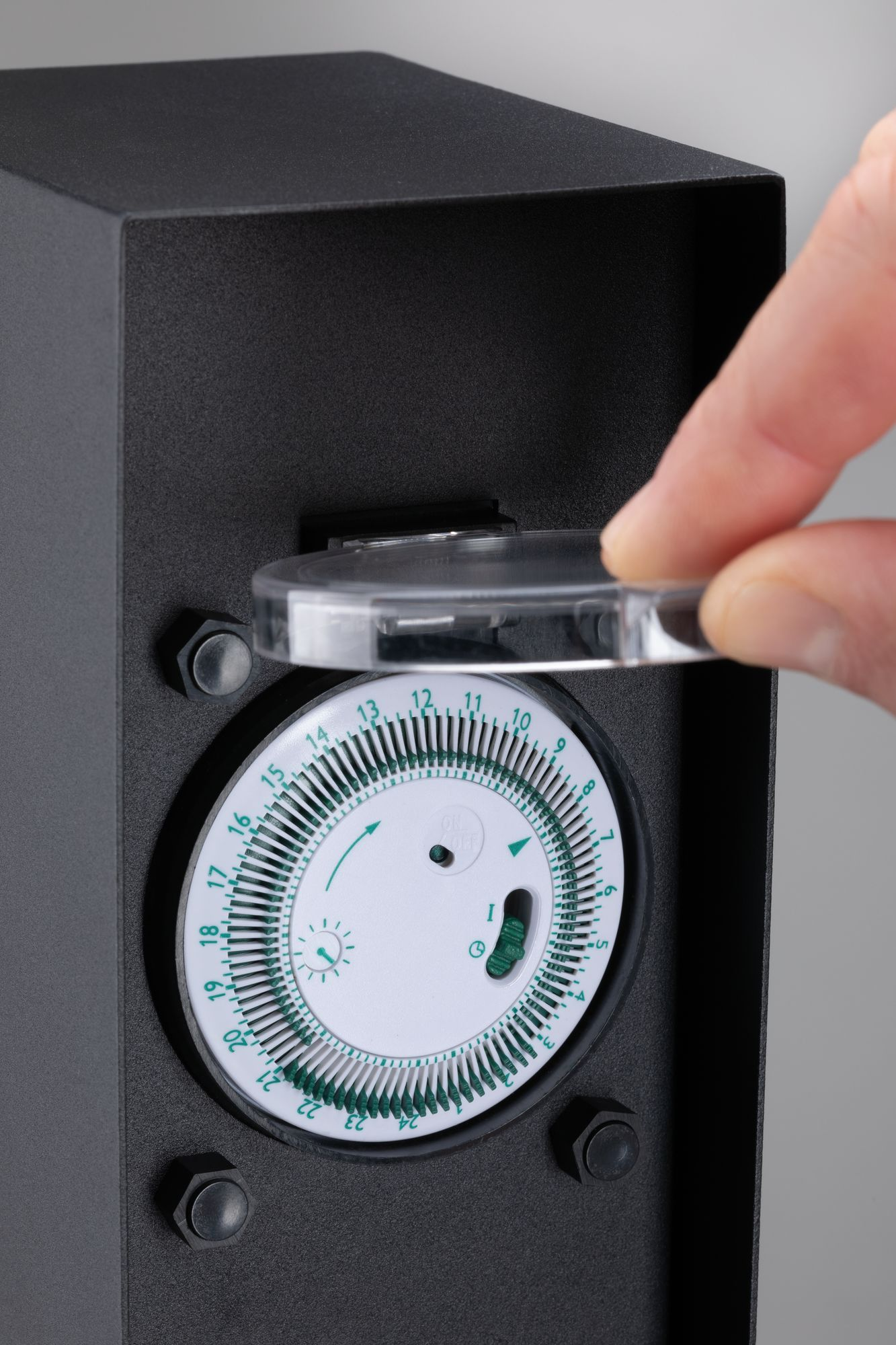 Ręka otwiera małą, przezroczystą pokrywę czarnego timera elektrycznego, odsłaniając tarczę.