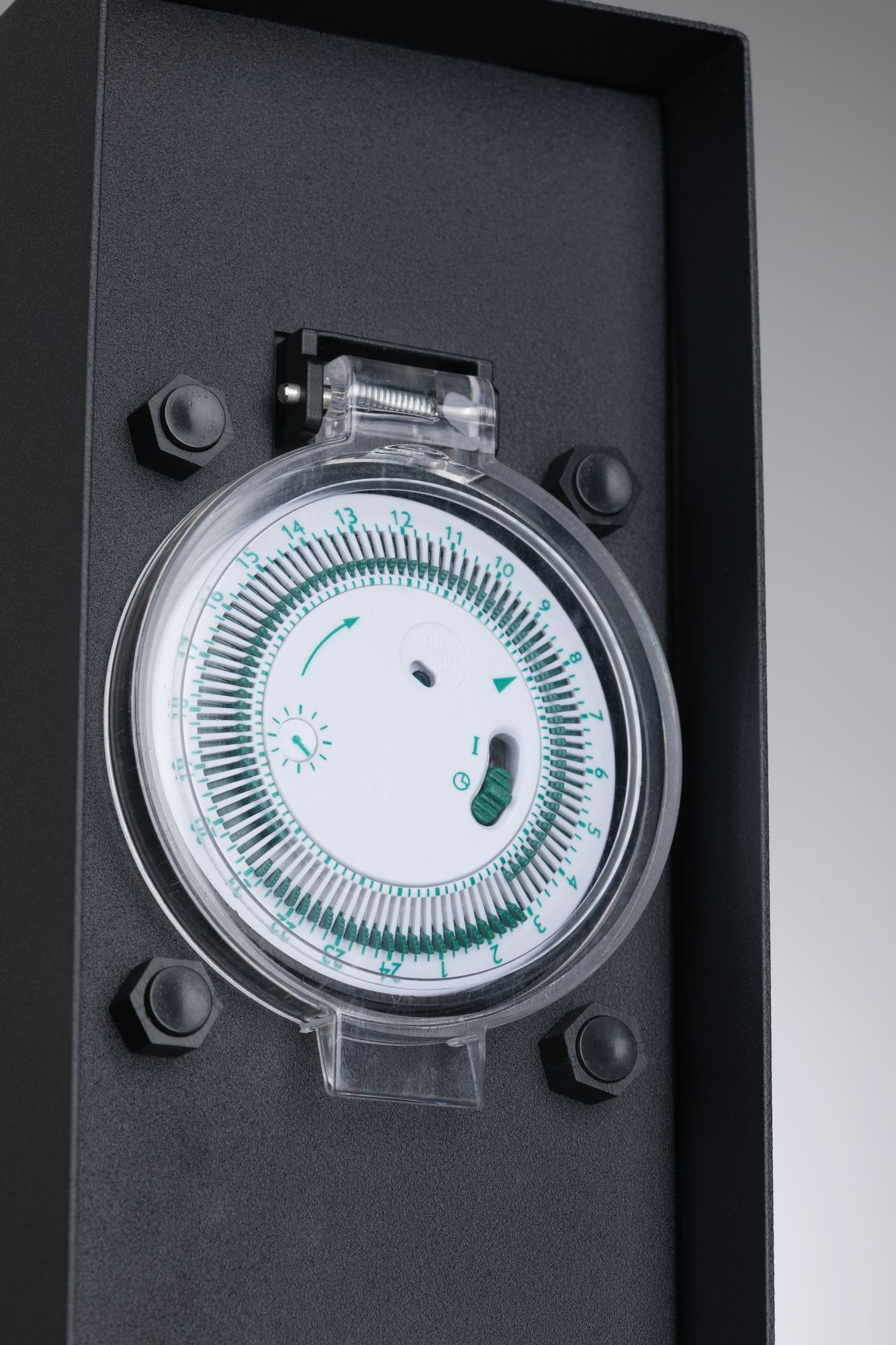 Czarny timer elektryczny z przezroczystą tarczą i zielonymi wskaźnikami. Zamontowany na czarnej powierzchni.