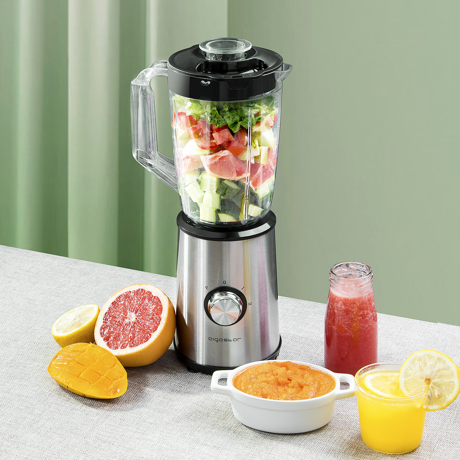 Mixer mit gehacktem Gemüse, Saftglas, Püreeschale und geschnittenen Früchten auf einem Tisch.