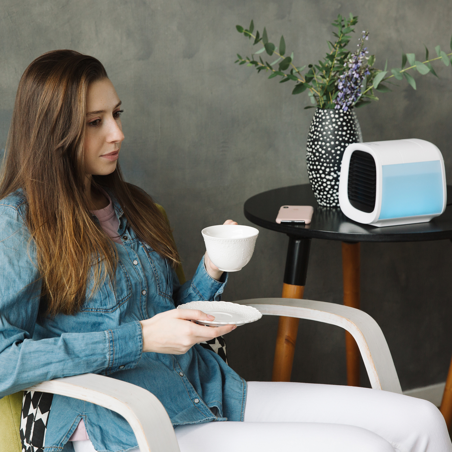 Frau sitzt, hält eine Tasse und Untertasse. Ein kleiner Tisch hat eine Vase mit Blumen und einen kleinen Luftkühler.