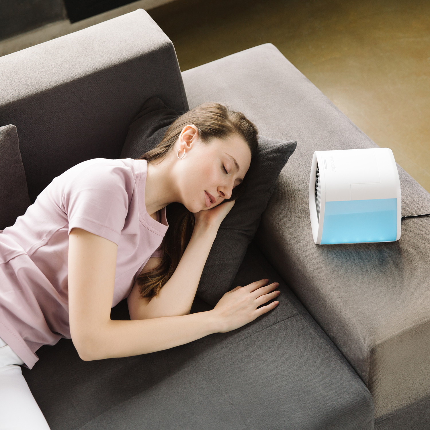 Eine Frau schläft auf einem Sofa neben einem kleinen, weißen Luftkühler mit blauem Licht.