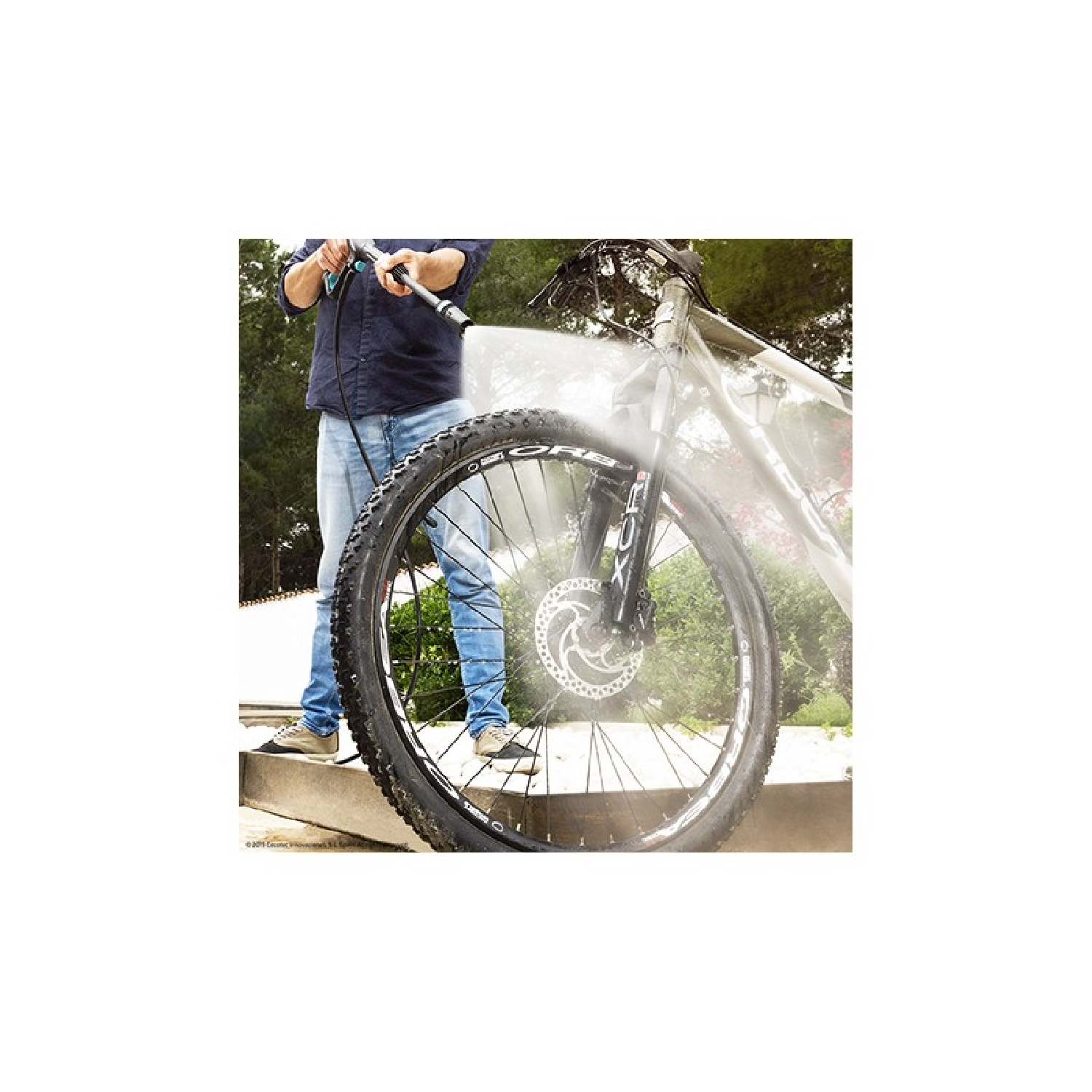 Persona lavando una bicicleta con una hidrolavadora. El agua rocía la rueda delantera. La escena es al aire libre.