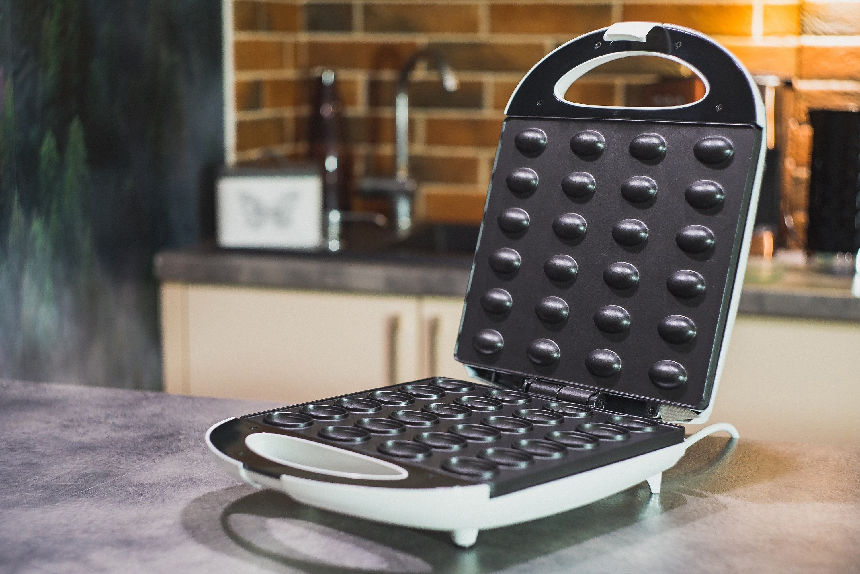 Otwarta gofrownica na blacie. Tło: zlewozmywak i ceglana ściana.