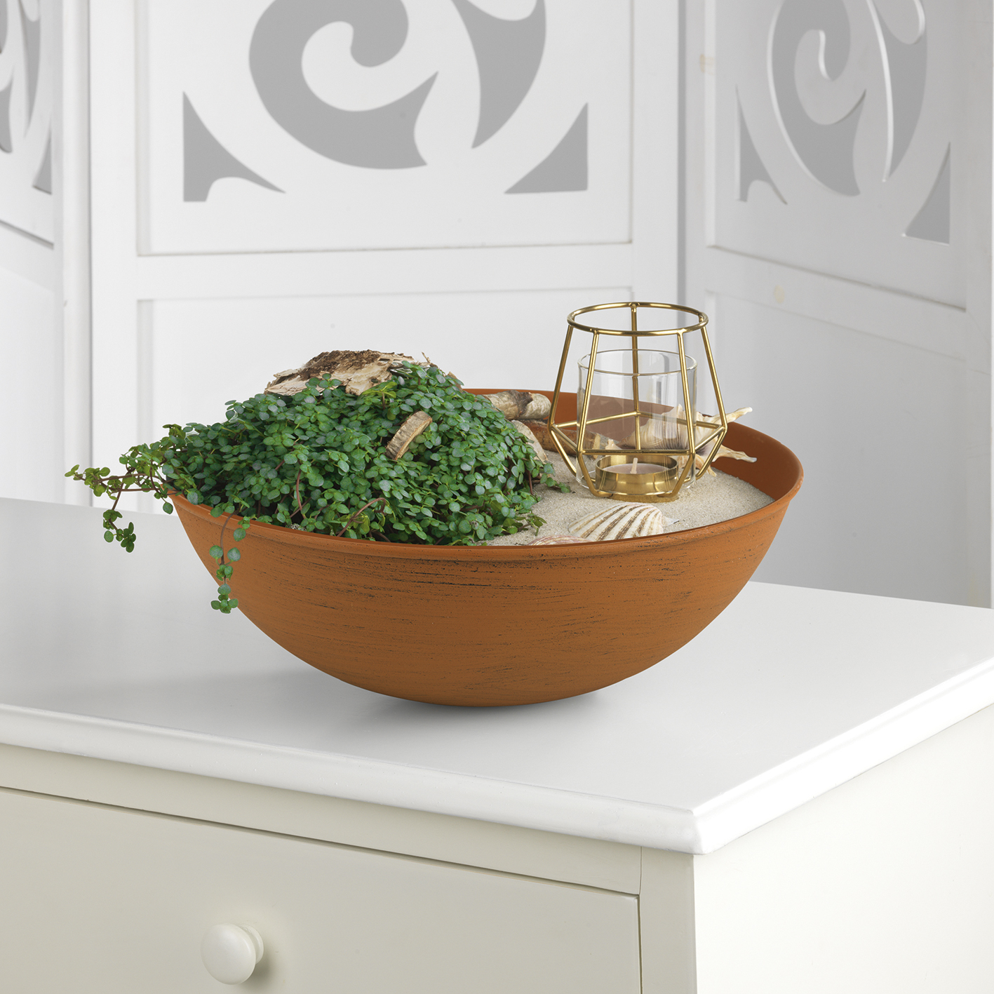 Terrakotta-Schale mit Pflanzen, Sand, Muscheln und einem Kerzenhalter auf weißem Tisch.