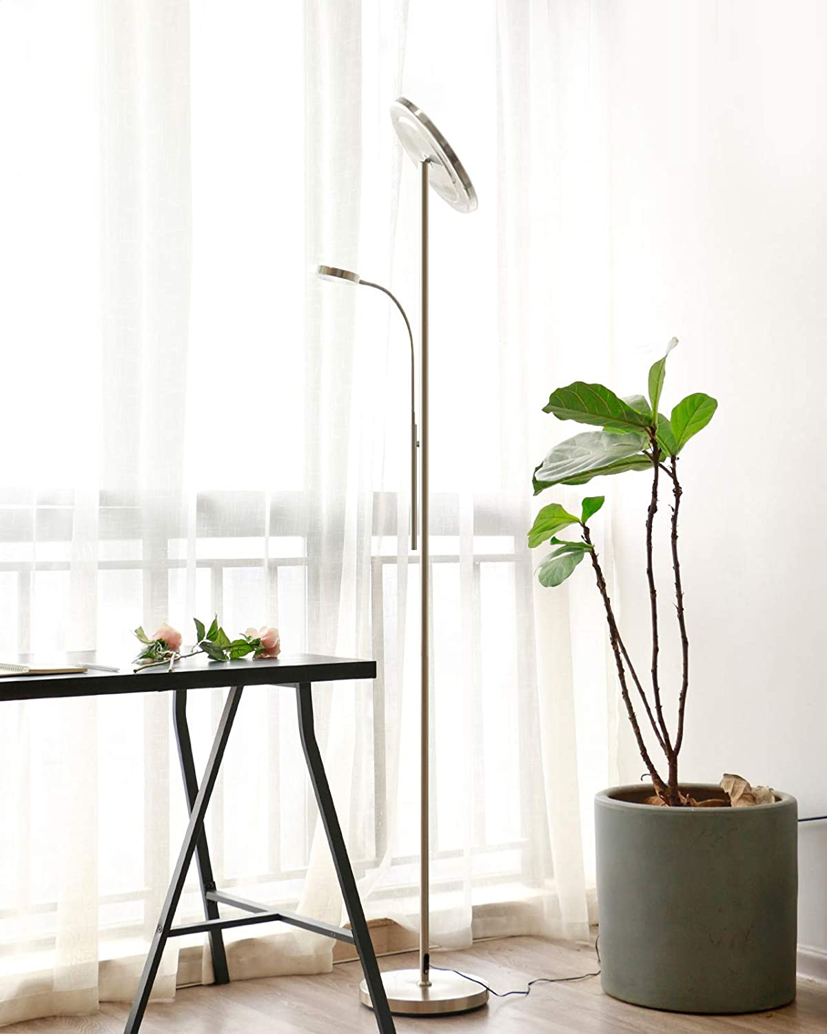 Hohe, moderne Stehlampe mit zwei Lichtern und einer Pflanze in einem grauen Topf. Fenster mit Vorhängen dahinter.