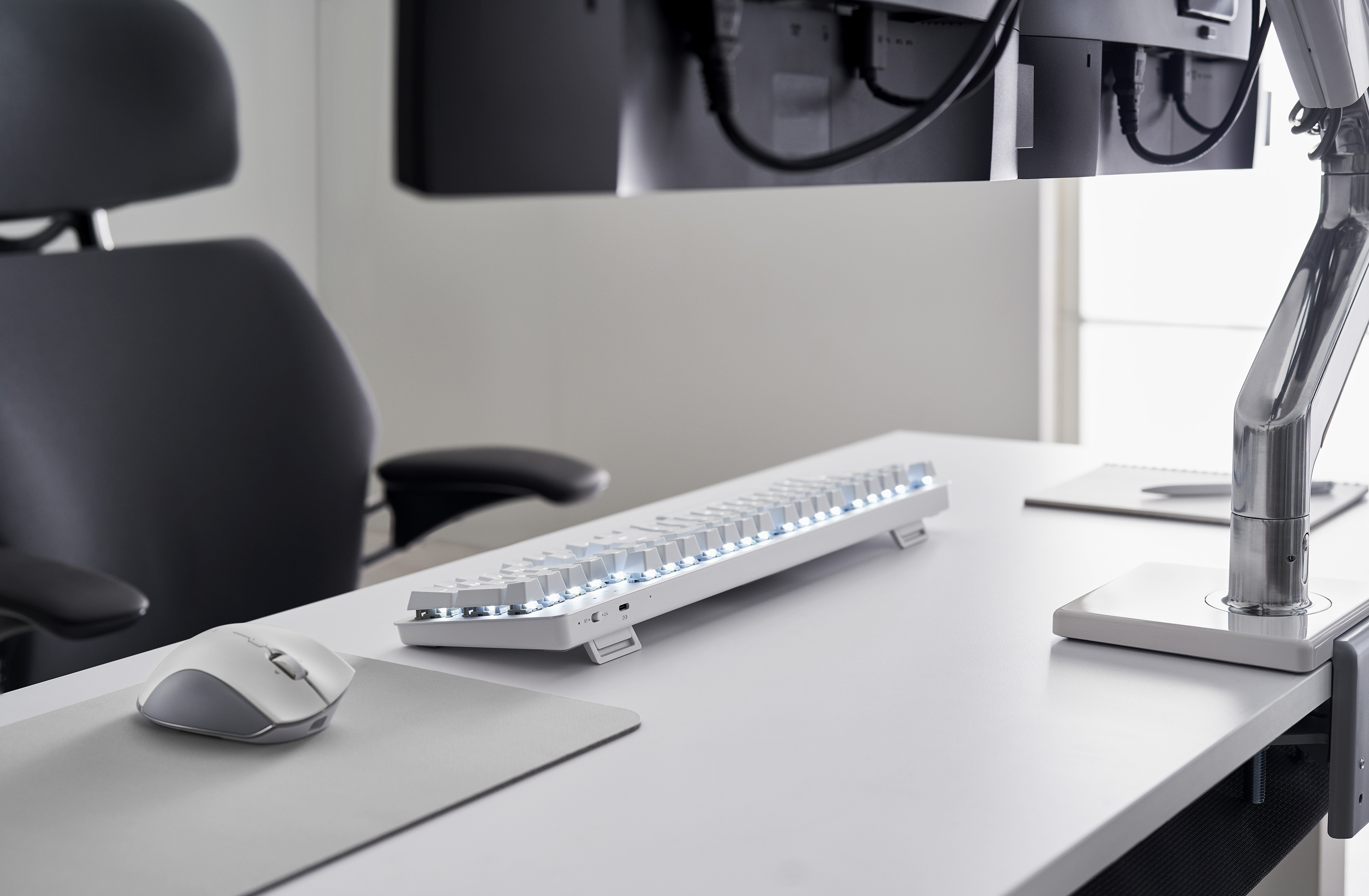 Biała klawiatura ze światłami na białym biurku. Szara mysz i stojak na monitor również są obecne.