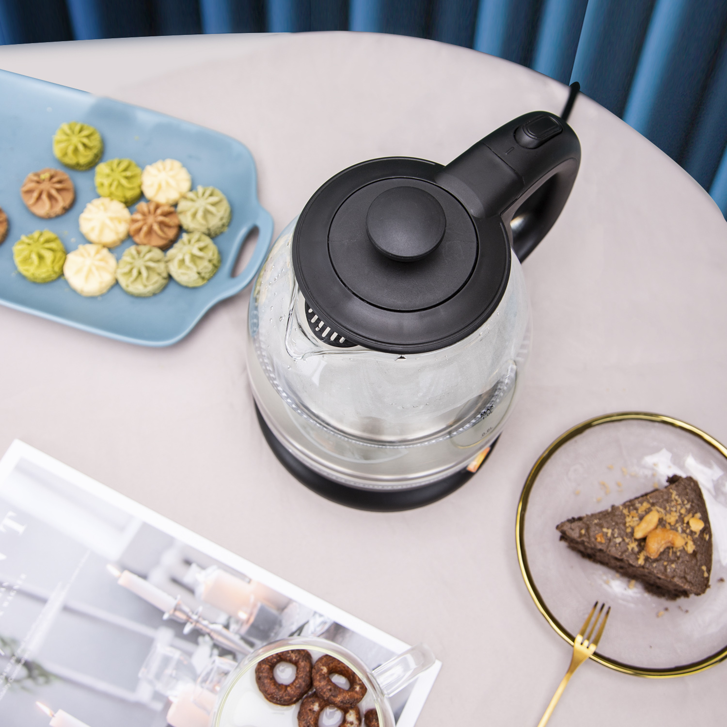 Glaskessel mit schwarzem Griff und Deckel auf Tisch mit Keksen und Kuchen.