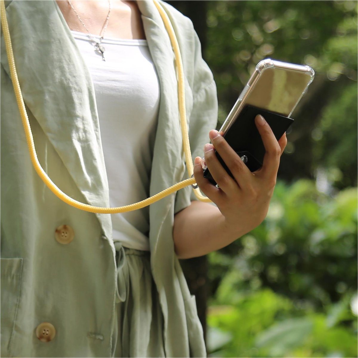 Donna che tiene il telefono in una custodia nera con cordino giallo. Lo sfondo è fogliame verde, sfocato.