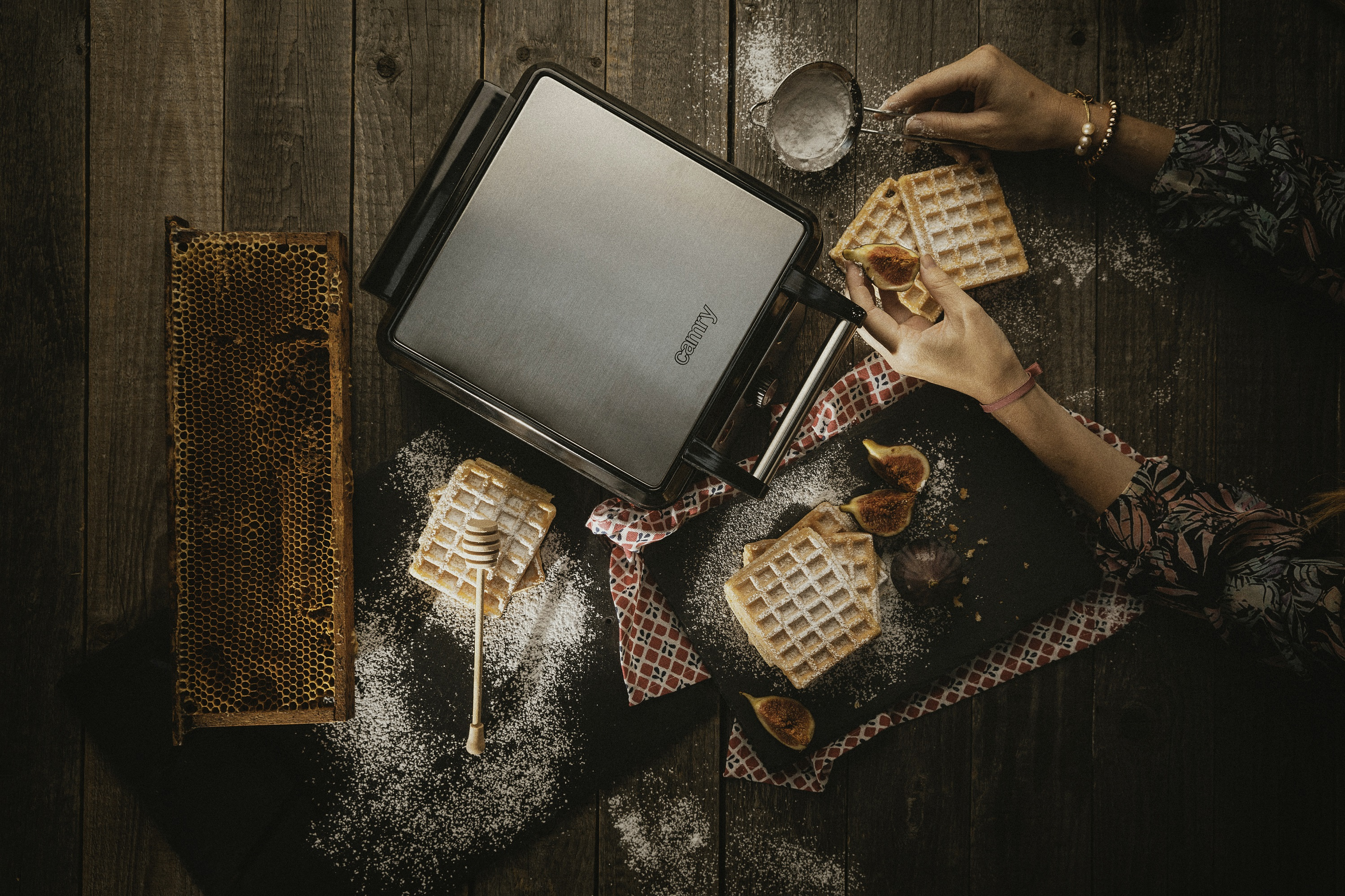 Kobieta przygotowuje gofry gofrownicą, miodem i figami na drewnianym stole.