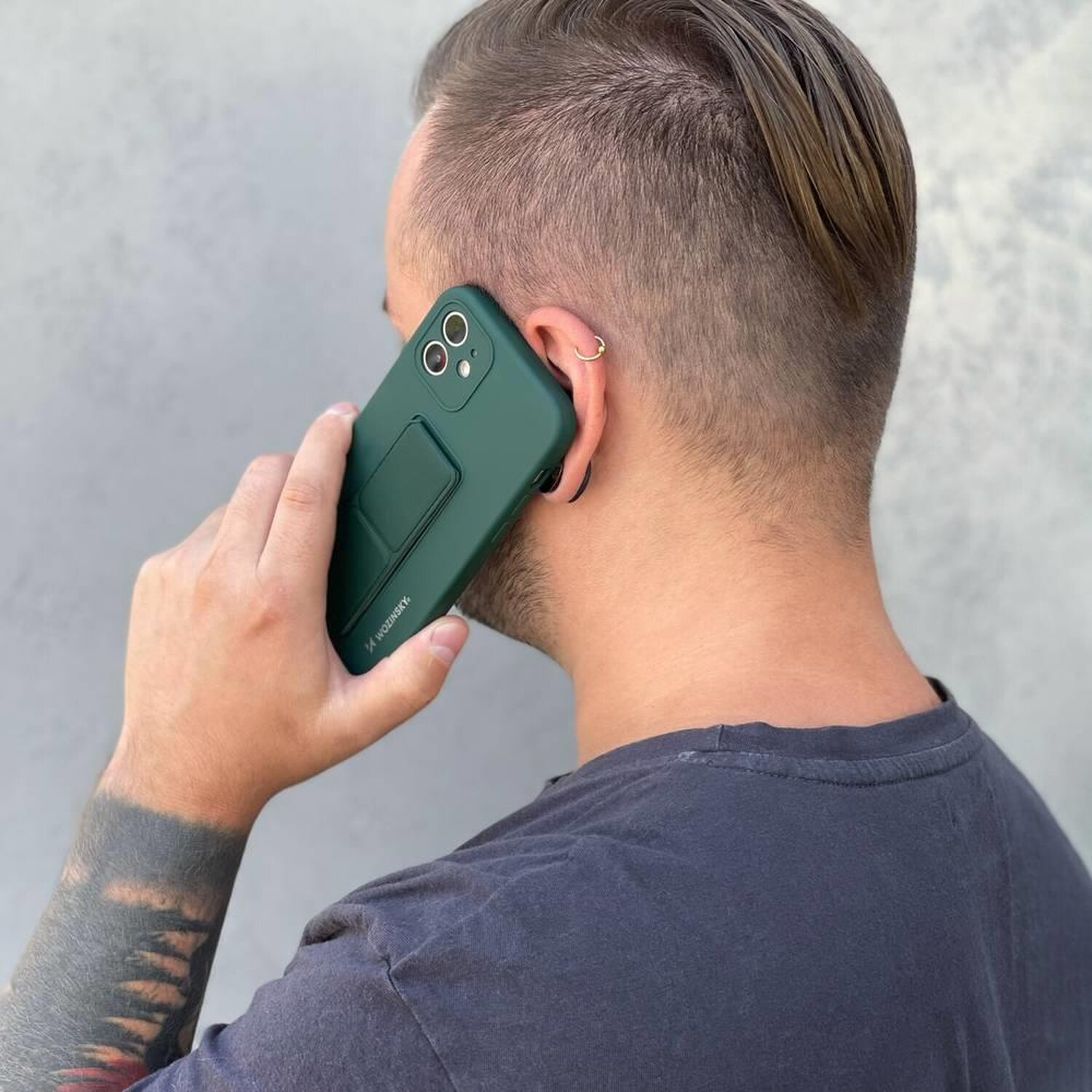 Ein Mann hält ein grünes Telefon an sein Ohr. Er hat ein Tattoo am linken Arm und ein Ohrpiercing.