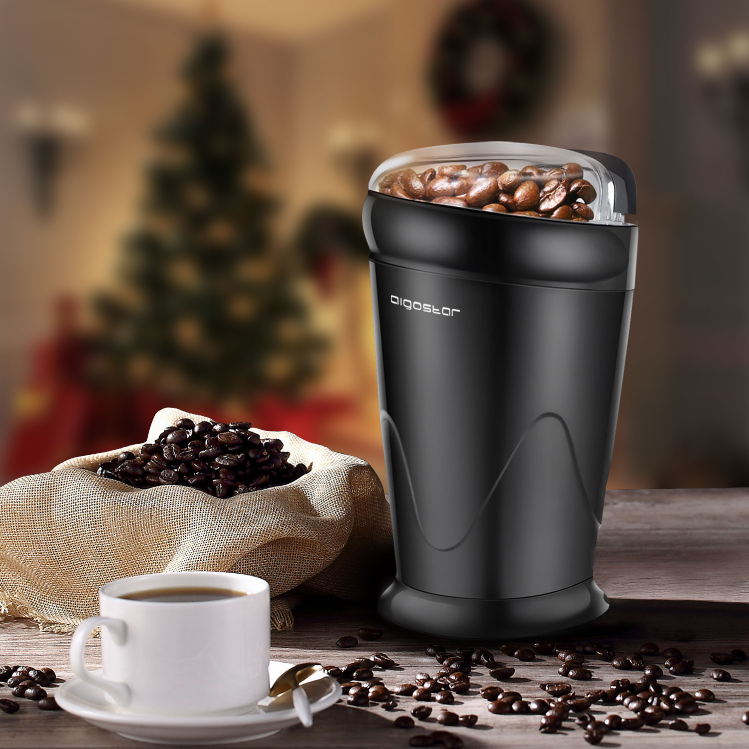 Molinillo de café negro con granos, taza de café. Árbol de Navidad al fondo.