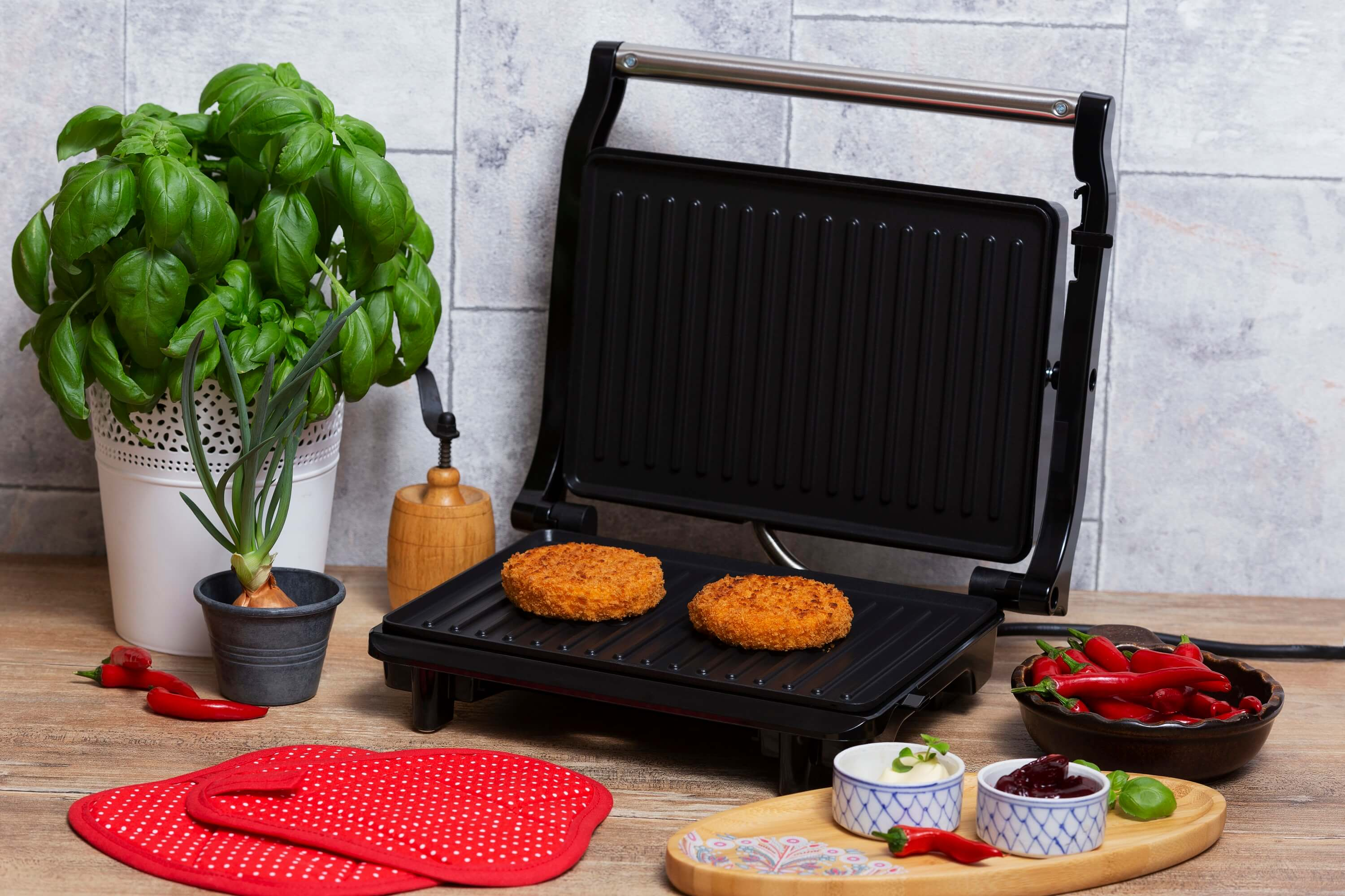 Otwarty grill z dwoma kotletami do burgerów, bazylią, papryką i przyprawami na drewnianym stole.