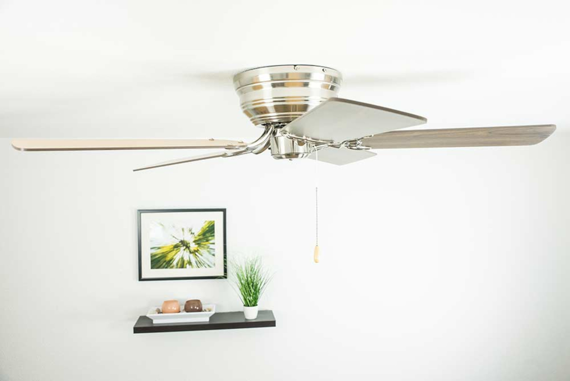 Ventilatore da soffitto con motore e pale argentate. Un quadro e una piccola mensola sono sul muro bianco.