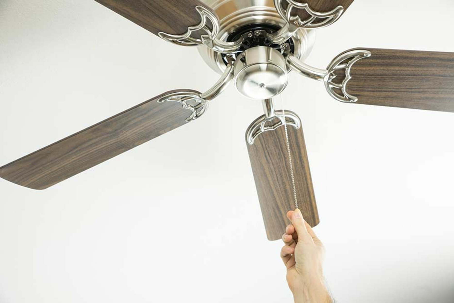 Mano che tira una catena per accendere un ventilatore a soffitto con pale in legno, soffitto bianco.