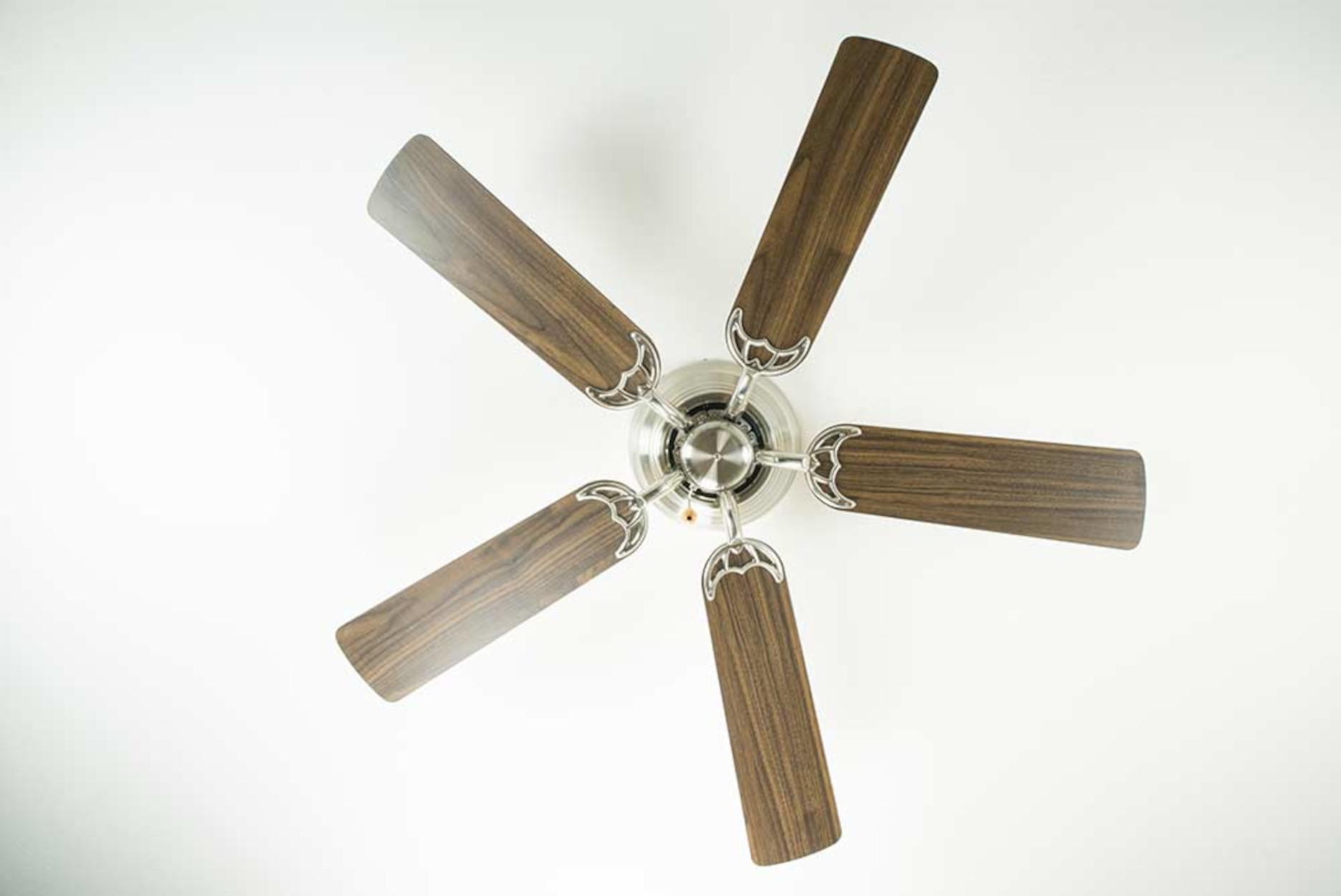 Ventilatore da soffitto con cinque pale in legno contro un soffitto bianco, visto dal basso.