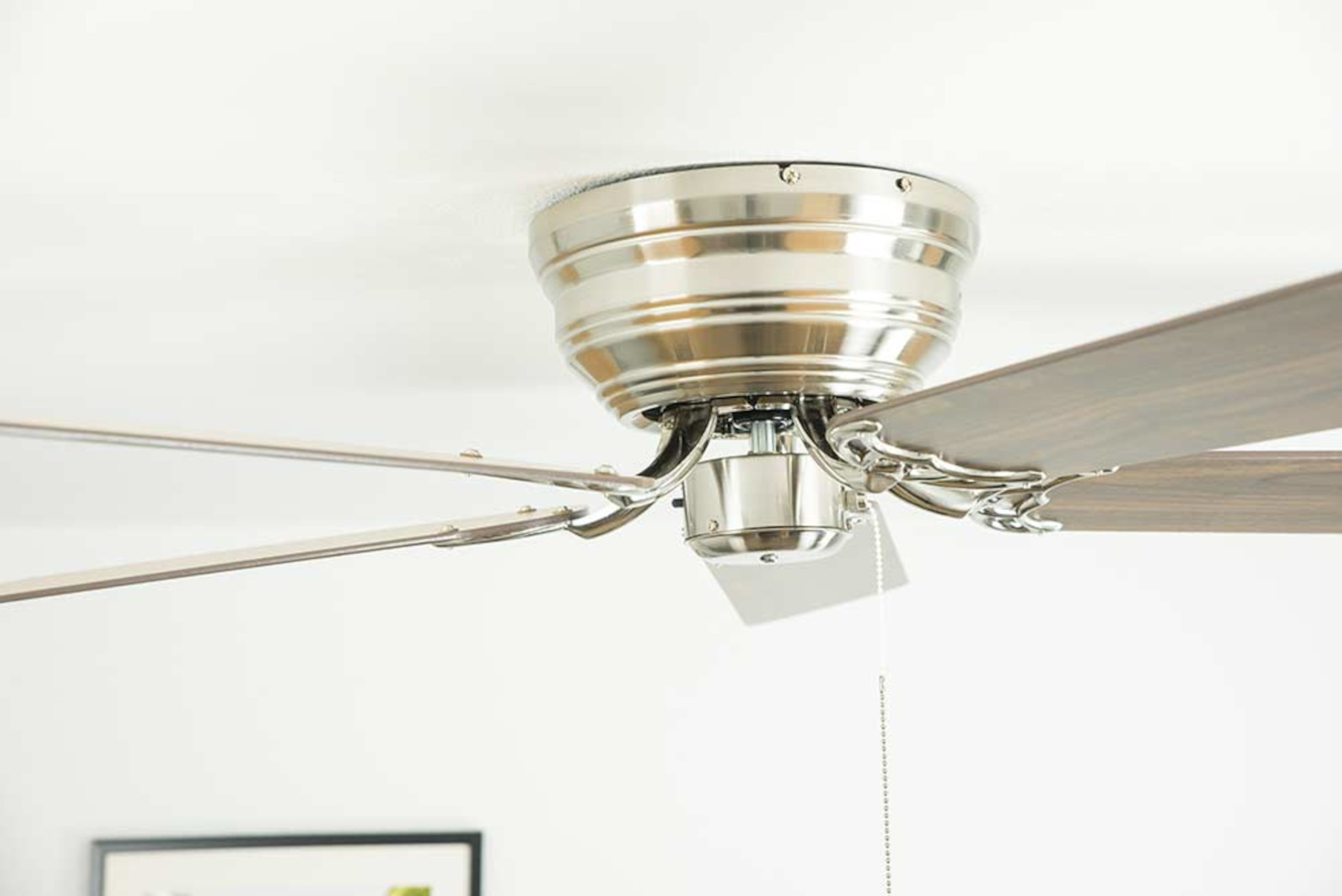 Un ventilatore da soffitto con cassa in metallo argentato e pale in legno è installato su un soffitto bianco.