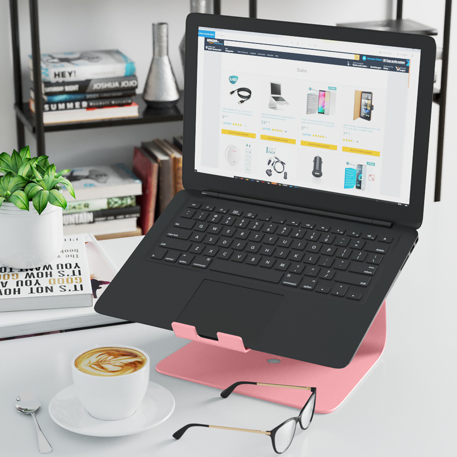 Ein Laptop auf einem rosa Ständer. Daneben stehen eine Kaffeetasse und eine Brille sowie Bücher.