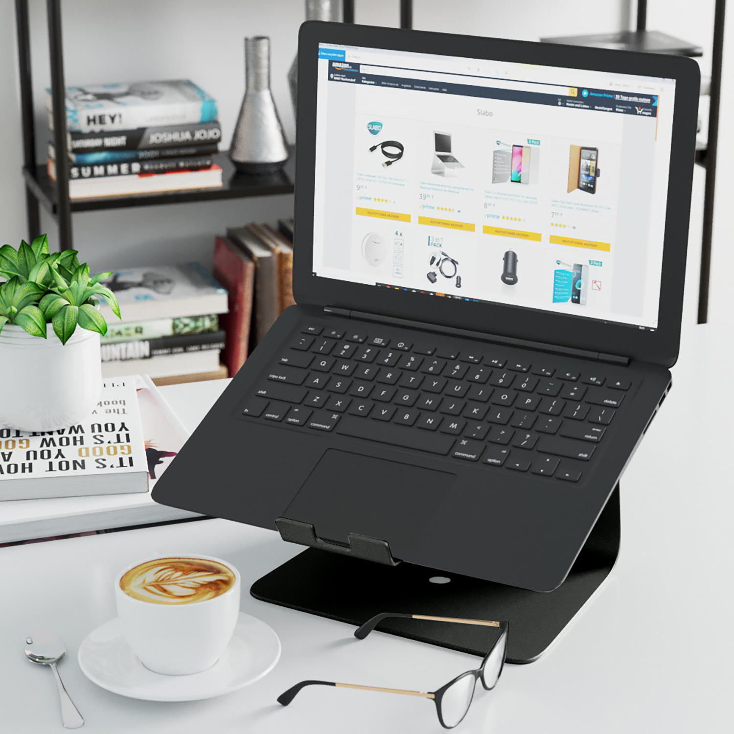 Schwarzer Laptop auf Ständer, Kaffee, Brille und Bücher auf weißem Schreibtisch. Amazon-Website auf dem Bildschirm.