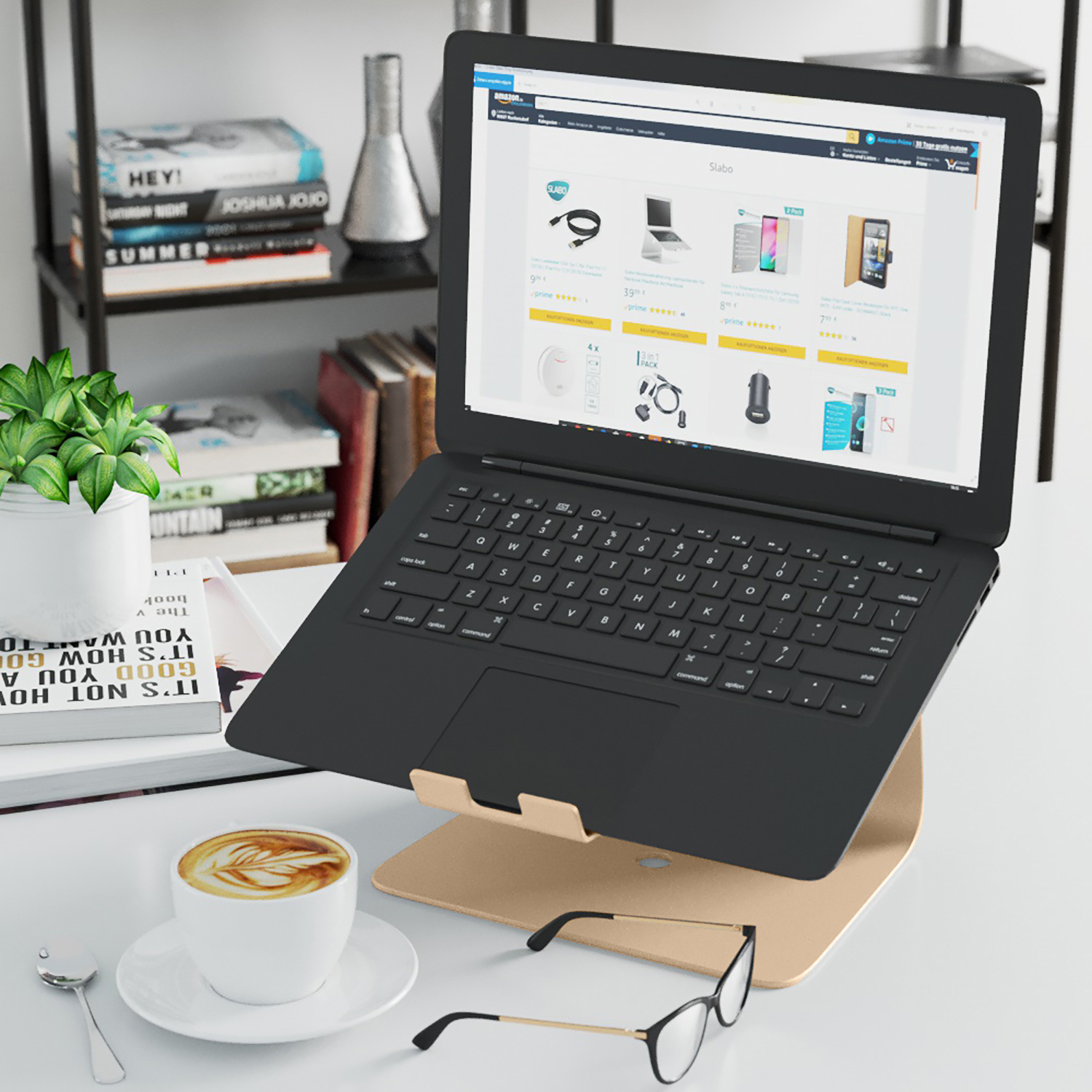 Ein Laptop auf einem goldenen Ständer. Eine Kaffeetasse, Brille und Bücher stehen auf dem Schreibtisch. Eine Pflanze im Hintergrund.