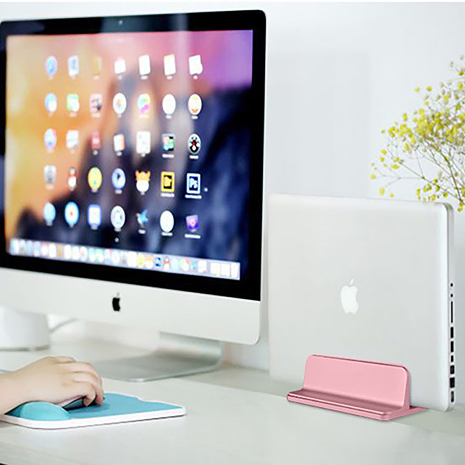 Computer-Setup: Monitor, Laptopständer, Maus. Weißer Schreibtisch. Gelbe Blumen im Hintergrund.
