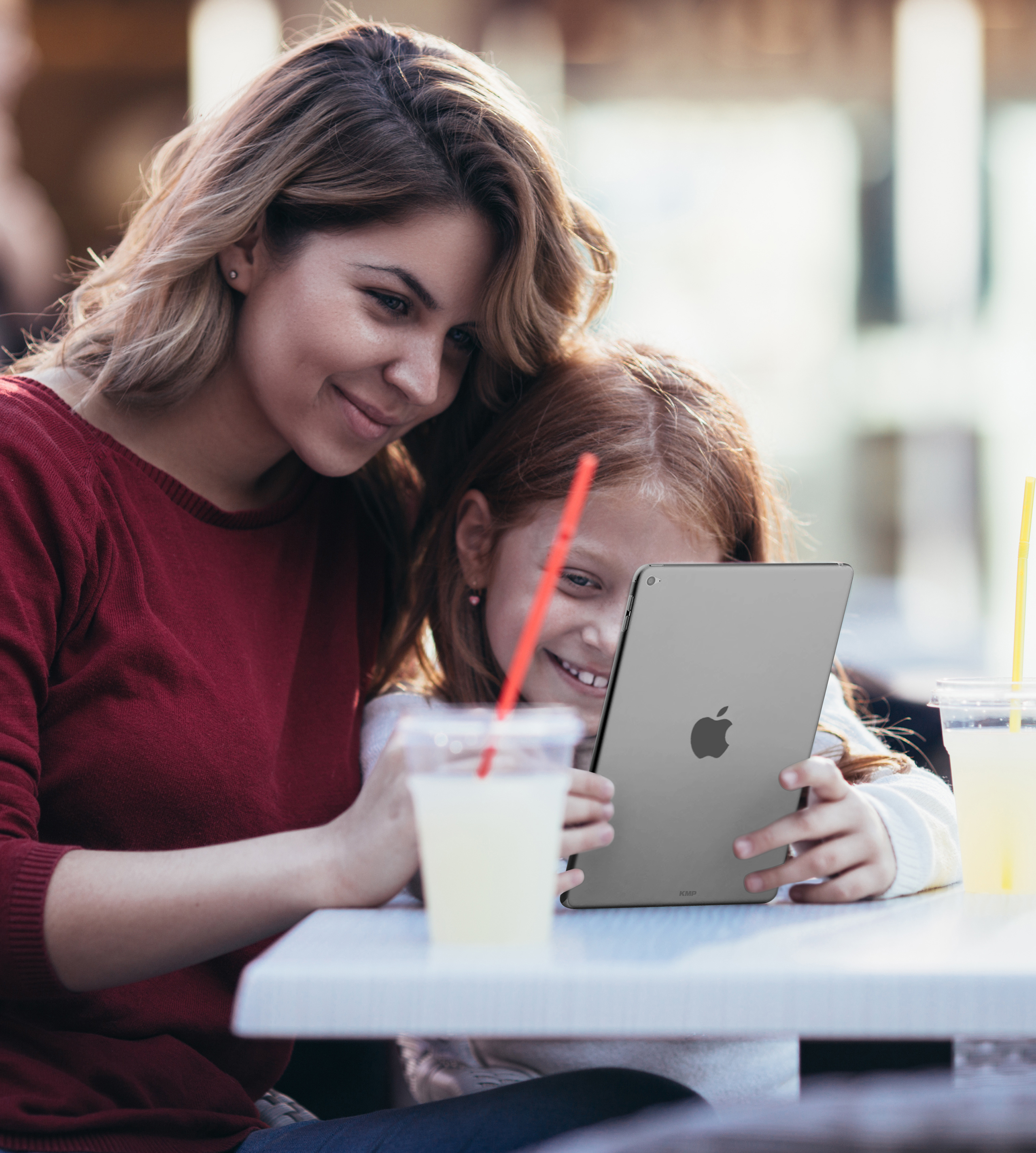 Eine Frau und ein Mädchen schauen auf ein graues iPad. Sie sitzen an einem Tisch mit Getränken.