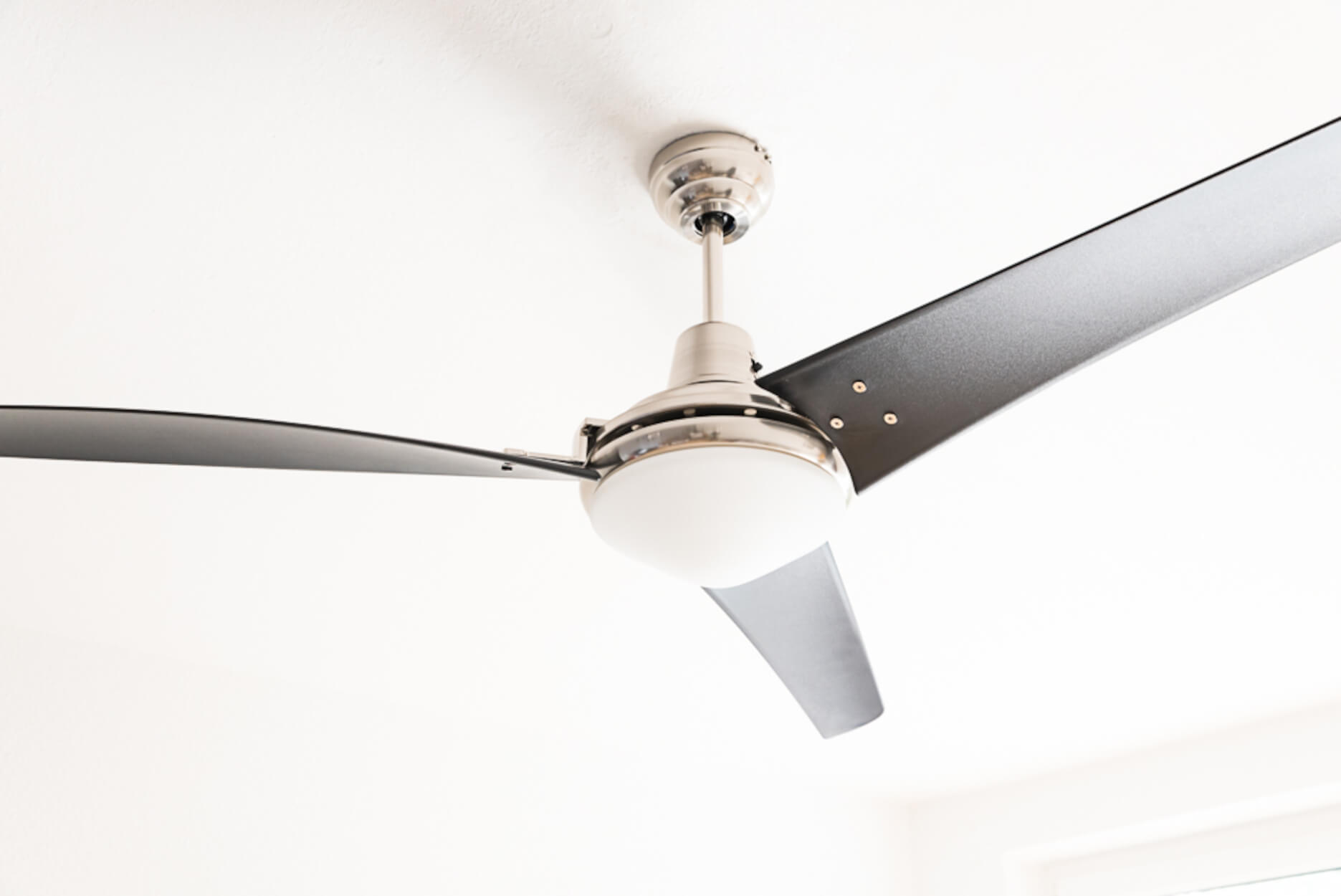 Un ventilatore da soffitto con tre pale nere e un apparecchio di illuminazione bianco. È montato su un soffitto bianco.
