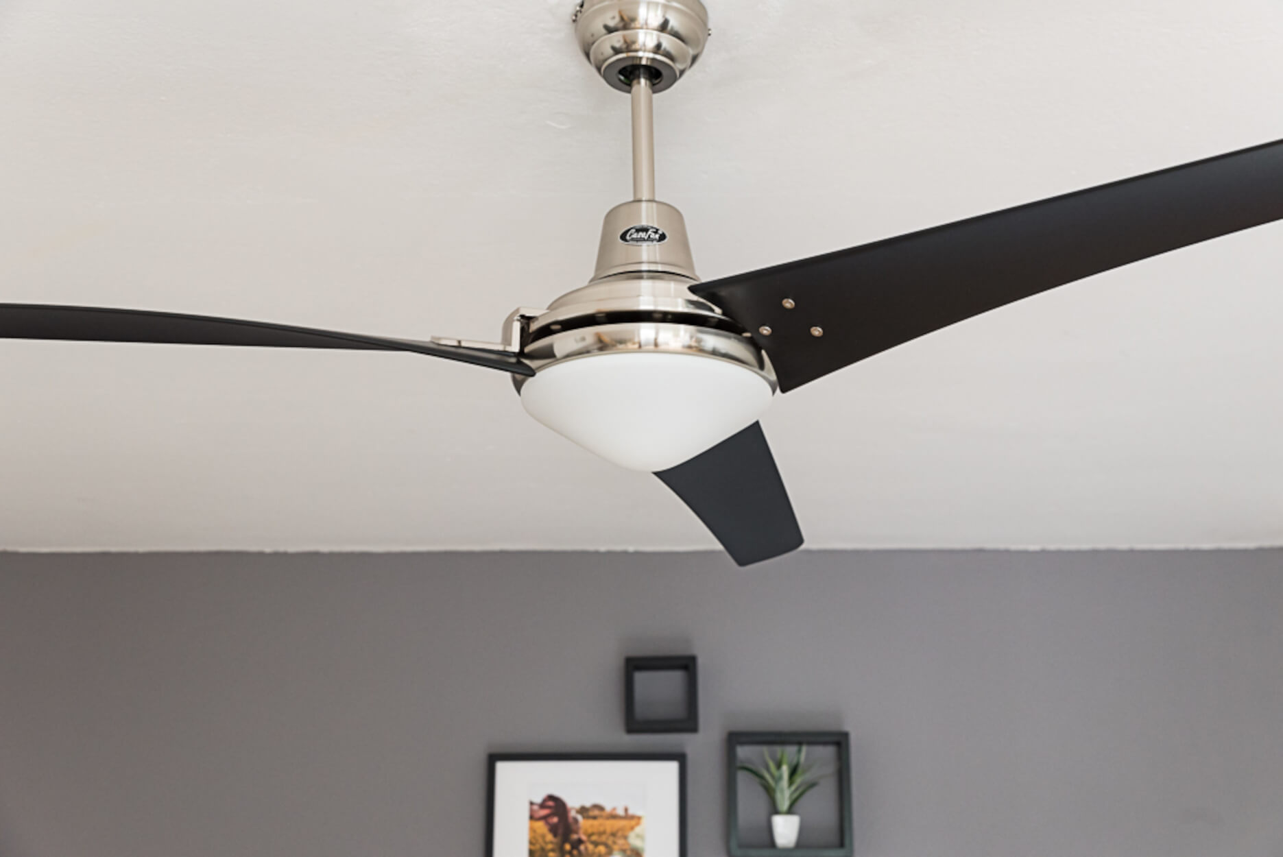 Ventilatore a soffitto. Base argentata, pale nere, luce bianca. Sfondo grigio.
