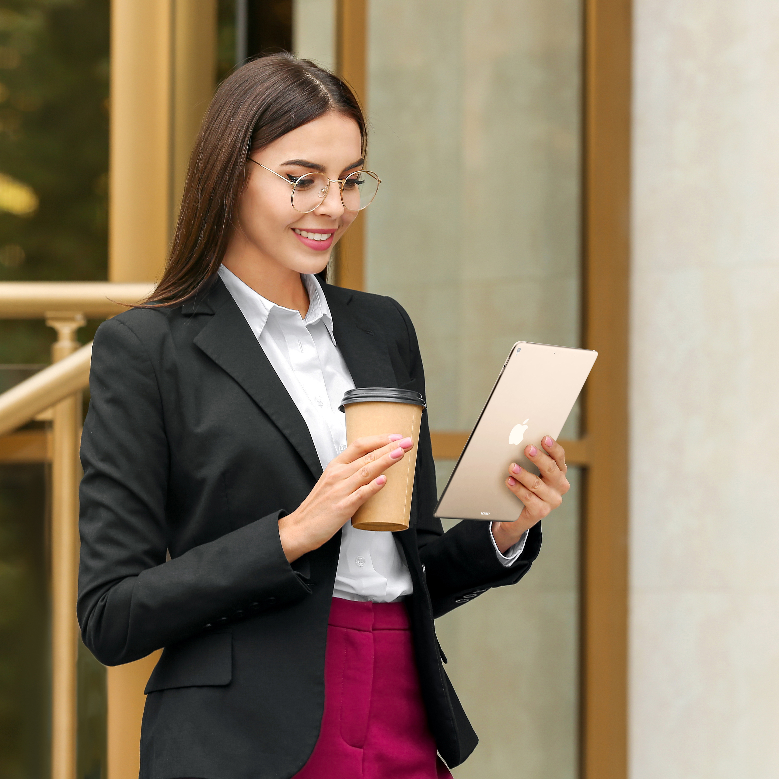 Eine Frau in einem schwarzen Blazer und einer rosa Hose benutzt ein Tablet und hält eine Kaffeetasse. Sie lächelt.