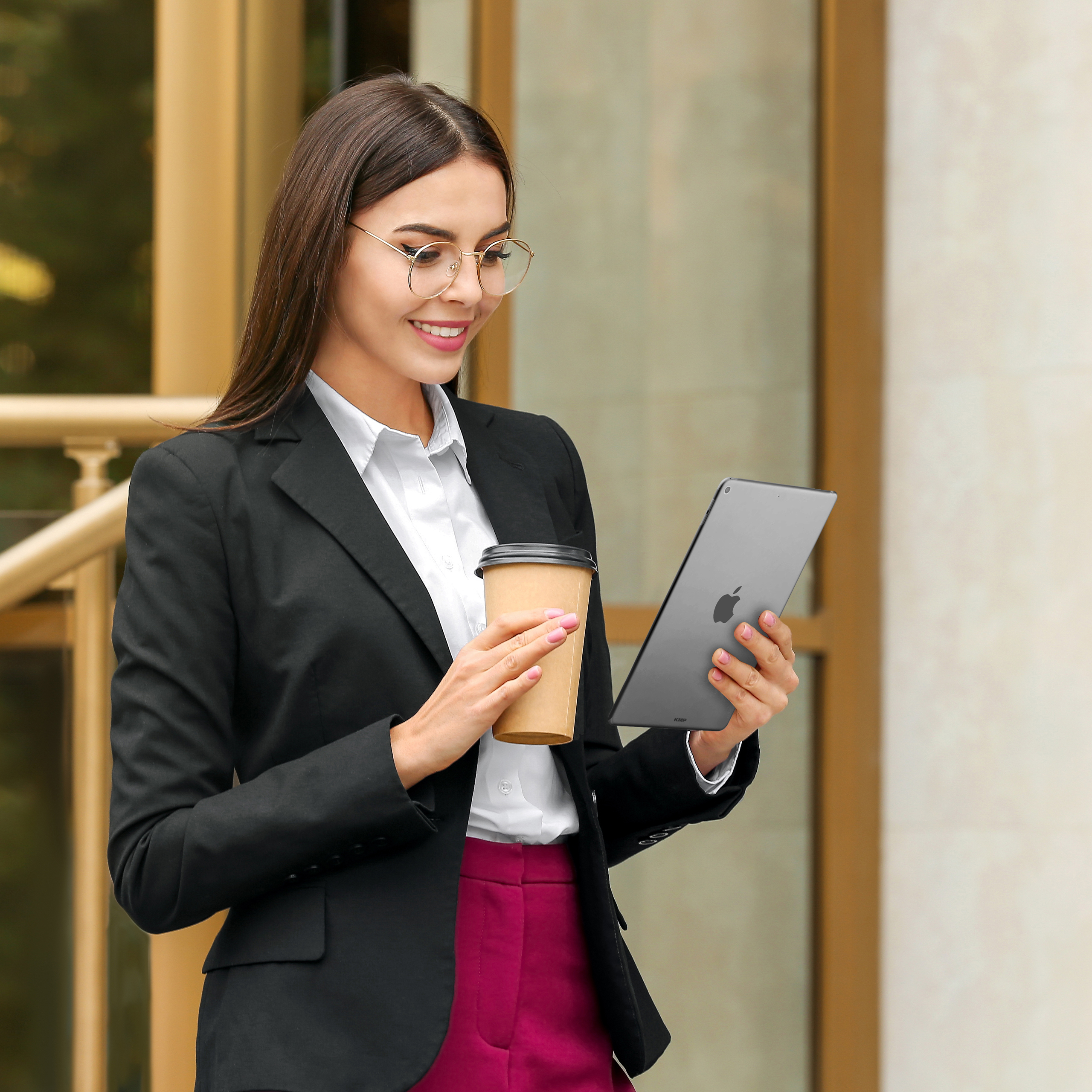 Frau mit Brille und Anzug hält Kaffeebecher und Tablet vor Gebäude.